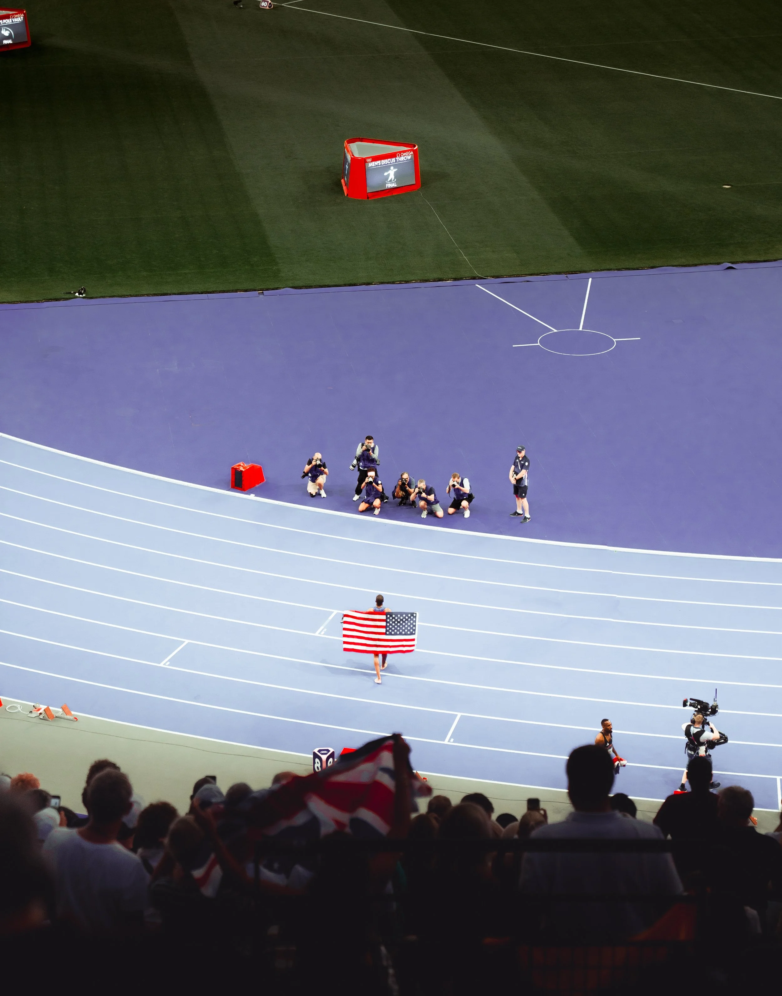 An athlete holding an American flag walks on a blue running track at a stadium, surrounded by photographers and spectators. The field and stadium seats are visible in the background.