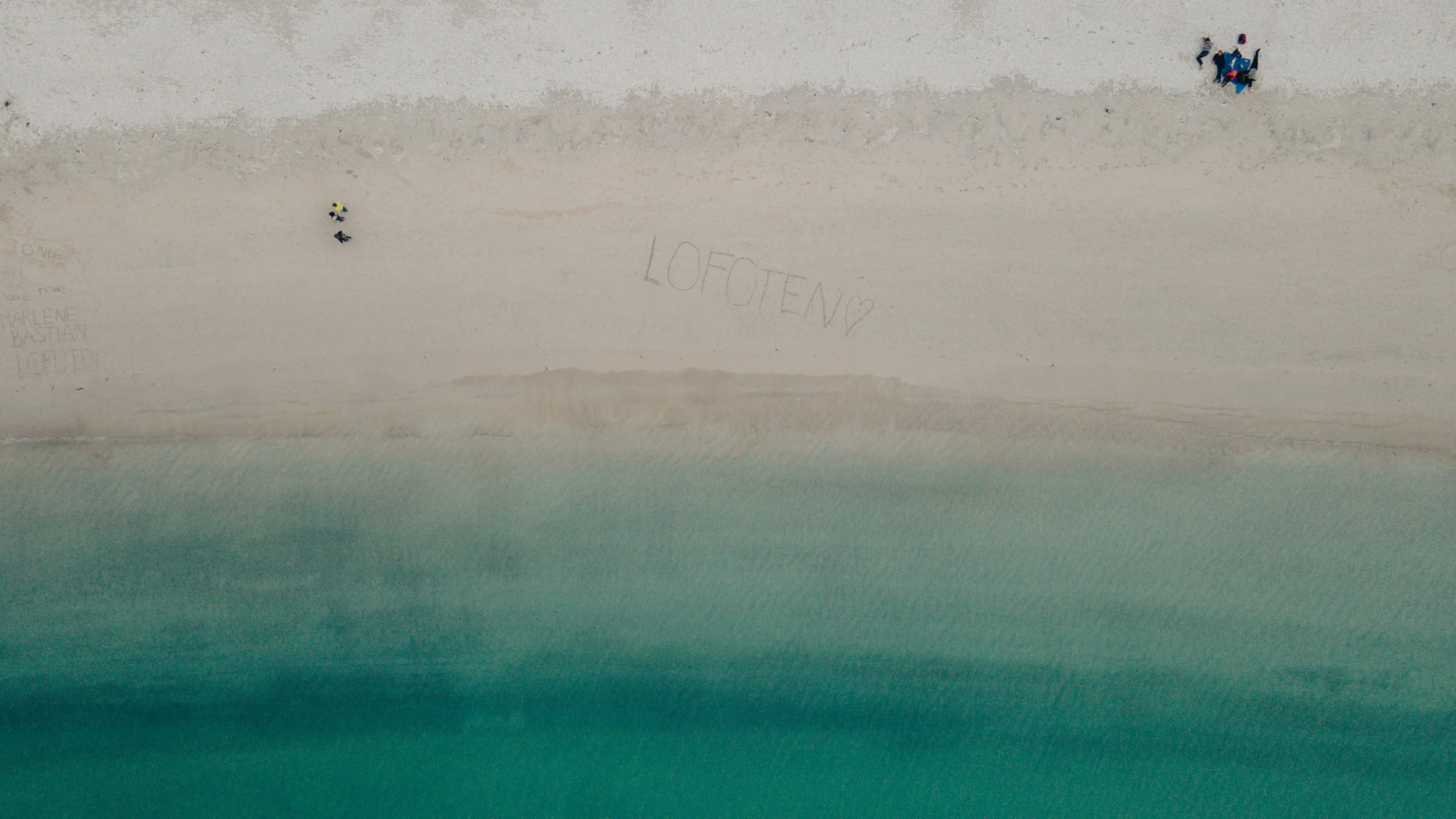 An aerial view of a beach with the words "LOFOTEN" and a heart drawn in the sand, and several people walking along the shoreline.