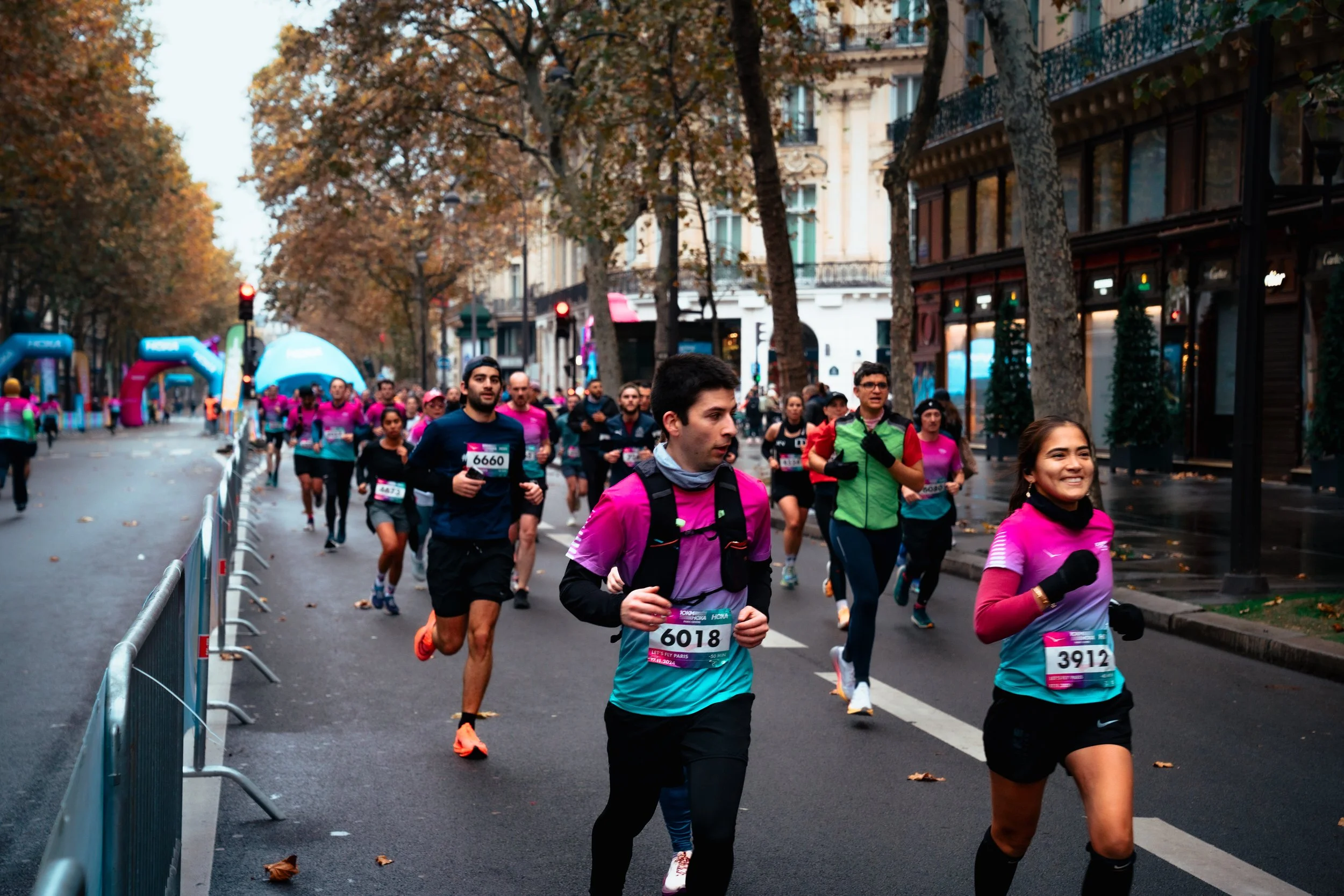 People participating in a marathon or running race on a city street with trees and buildings, some wearing pink shirts, race bibs, and athletic gear.