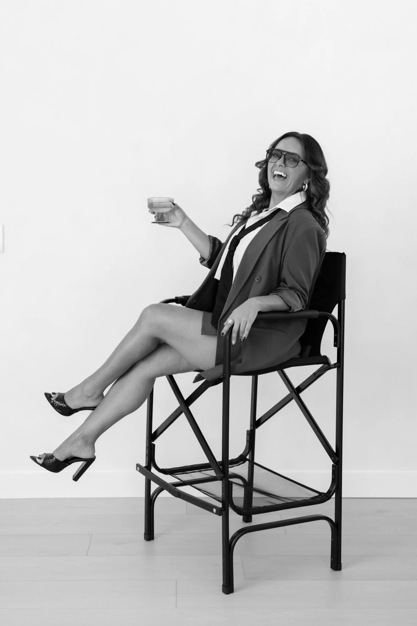 Black and white photo of a woman with long hair, glasses, and earrings, sitting on a folding chair with her legs crossed, holding a bowl in her right hand, wearing a blazer, blouse, skirt, and high heels, laughing against a plain background.