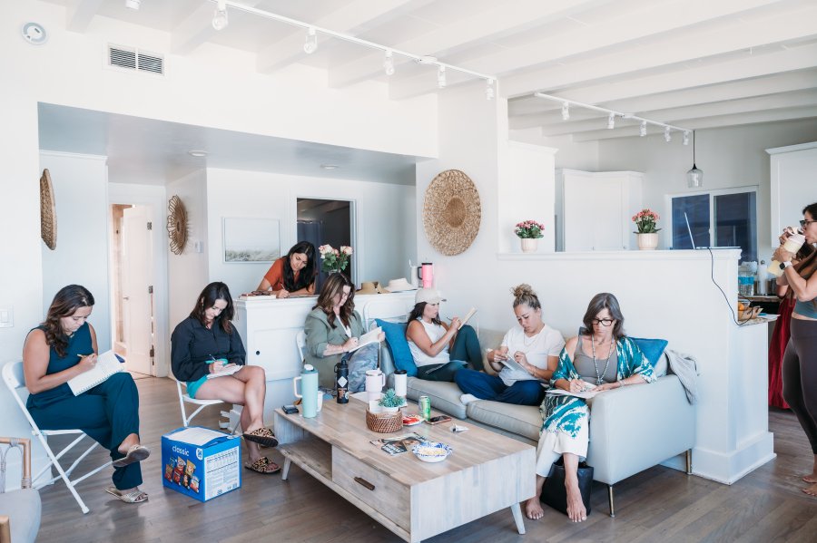 Bizz Bitch Community members participating in a creative indoor workshop, seated on chairs and sofas in a bright, modern space, writing in notebooks and engaging in focused collaborative activities.