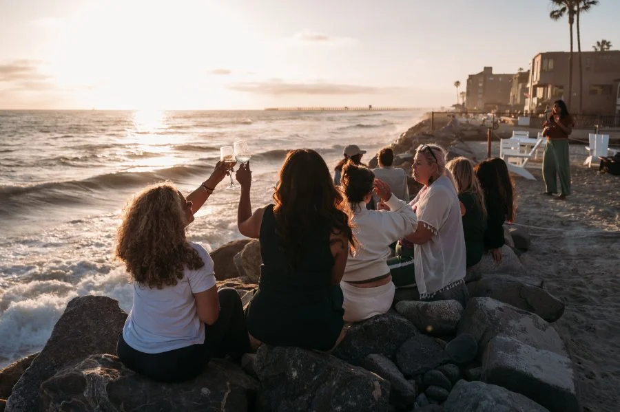 Bizz Bitch Community members celebrating on a rocky beach at sunset, raising glasses in a toast near the ocean waves, capturing a moment of connection and empowerment.