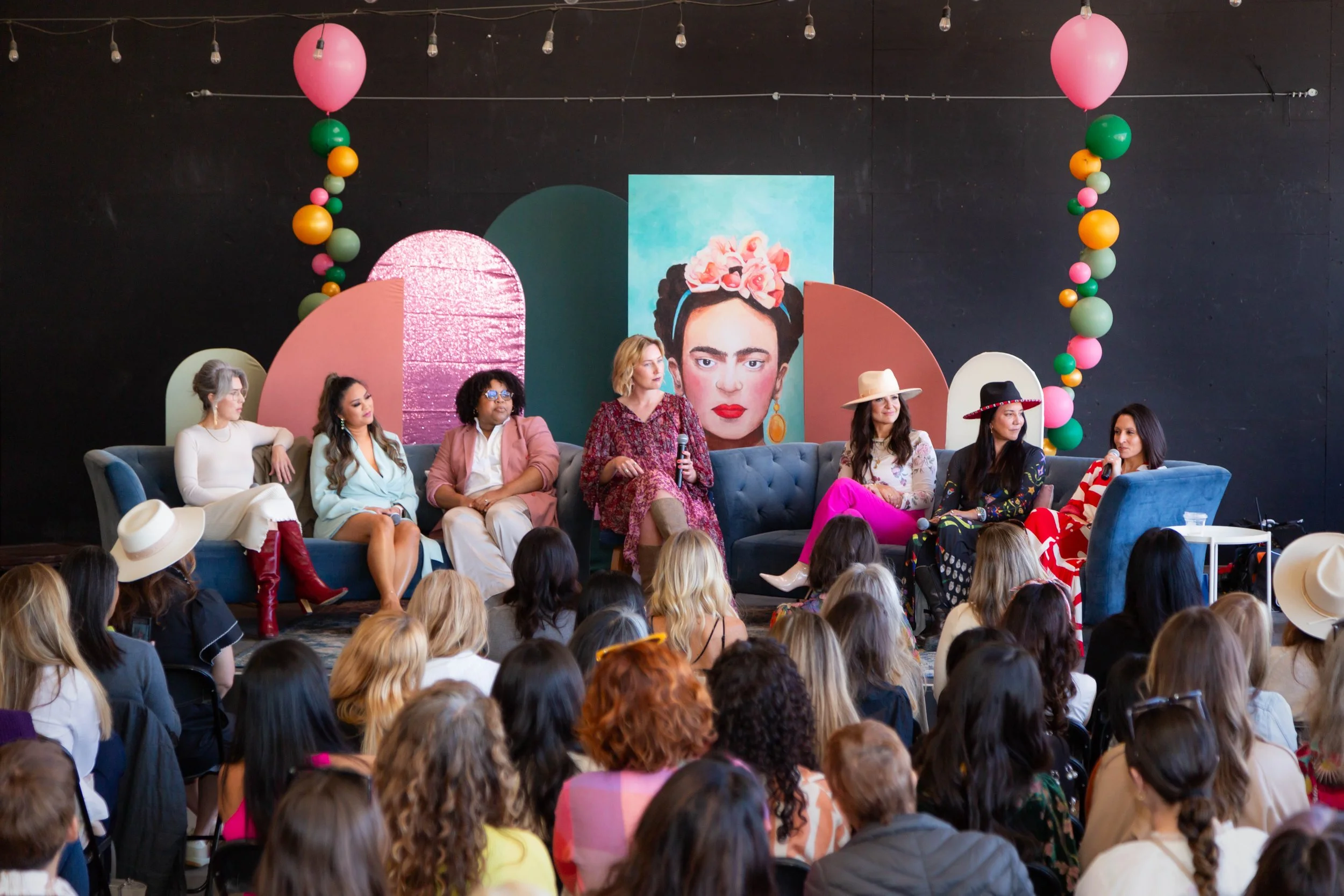 Empowered women panel discussion with a vibrant Frida Kahlo backdrop, lively balloon arch, and an engaged audience of women listening to diverse panelists at a creative event.