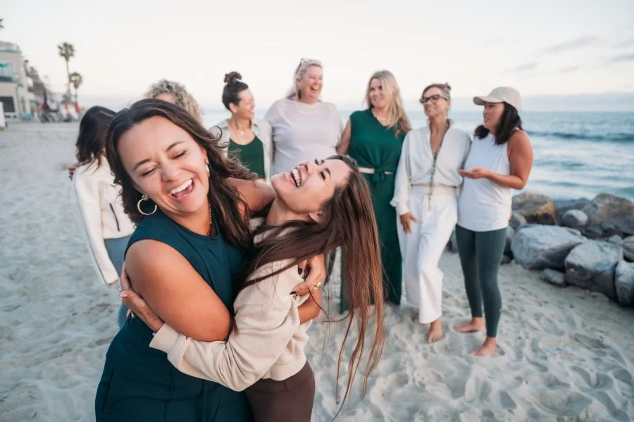 Jazmin Cornejo with Bizz Bitch Community members enjoying a beach gathering, laughing and embracing near the ocean. The group of women share a joyful moment of connection, friendship, and community by the shoreline.