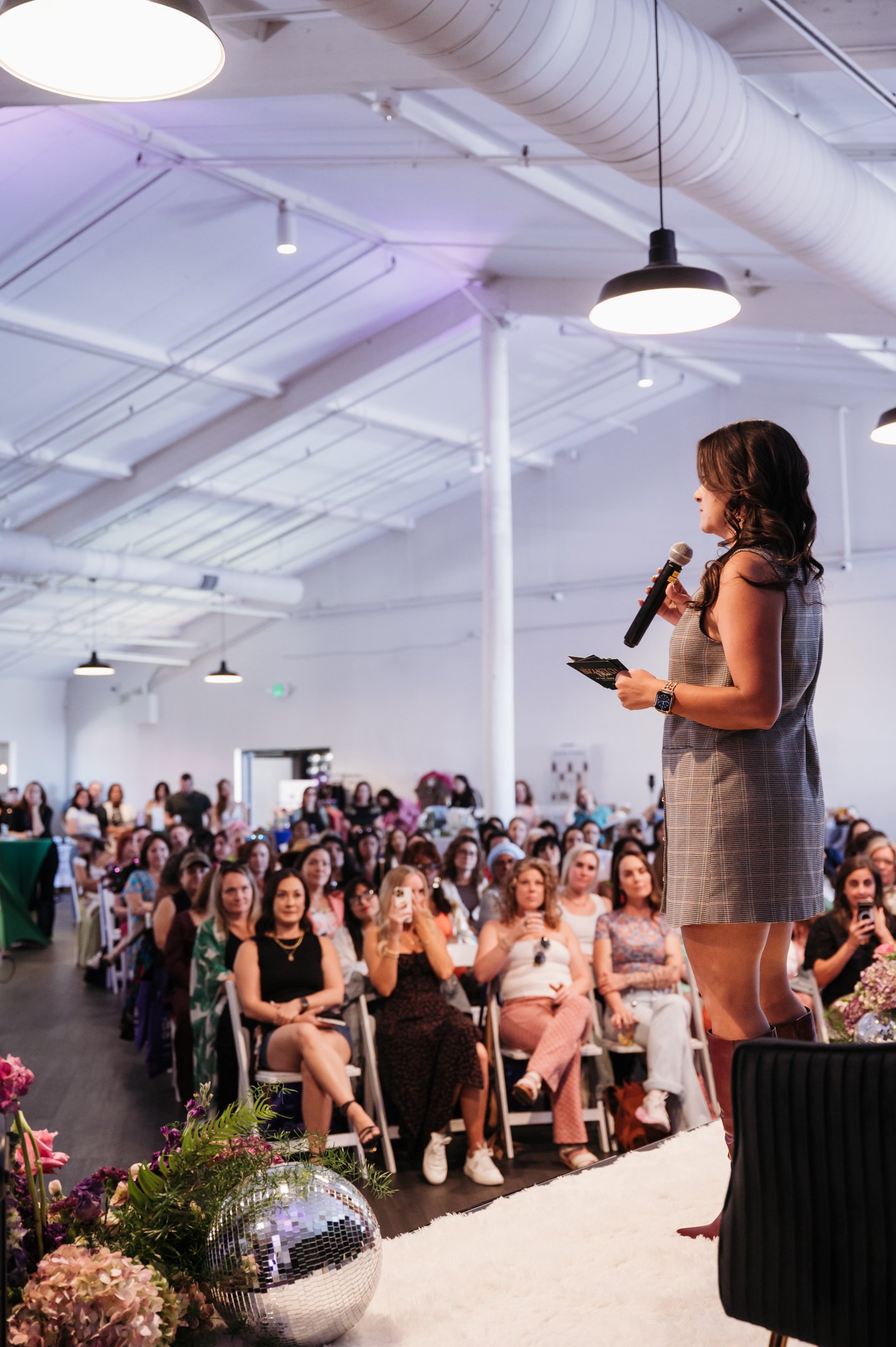 "Jazmin Cornejo speaking on stage at a business event, addressing a large audience with a microphone during an entrepreneurship and leadership presentation focused on community, networking, and women in business.