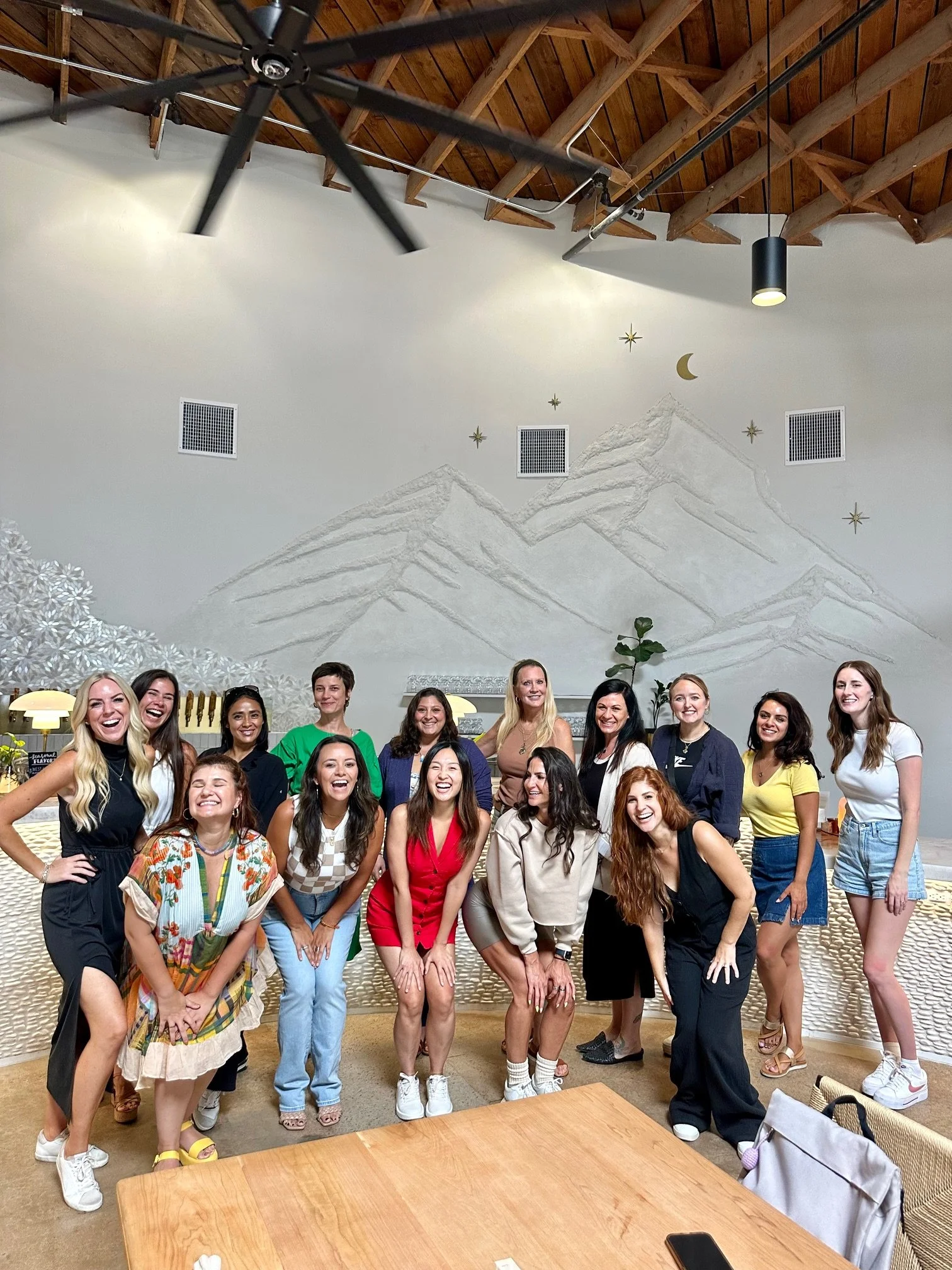 A group photo of 14 women smiling and posing together in a lively, modern setting. They are dressed in a variety of casual outfits, with a bright, cheerful atmosphere in the background, showcasing a sense of community and camaraderie.