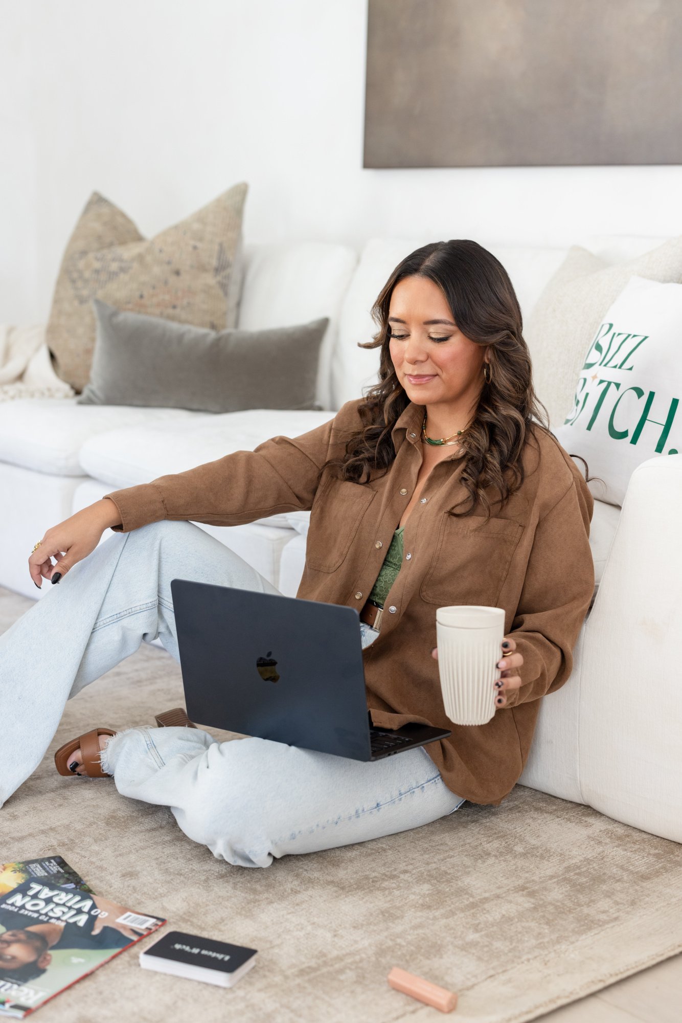 A woman sitting on the floor in a living room, using a laptop, holding a drink, with magazines and books around her.