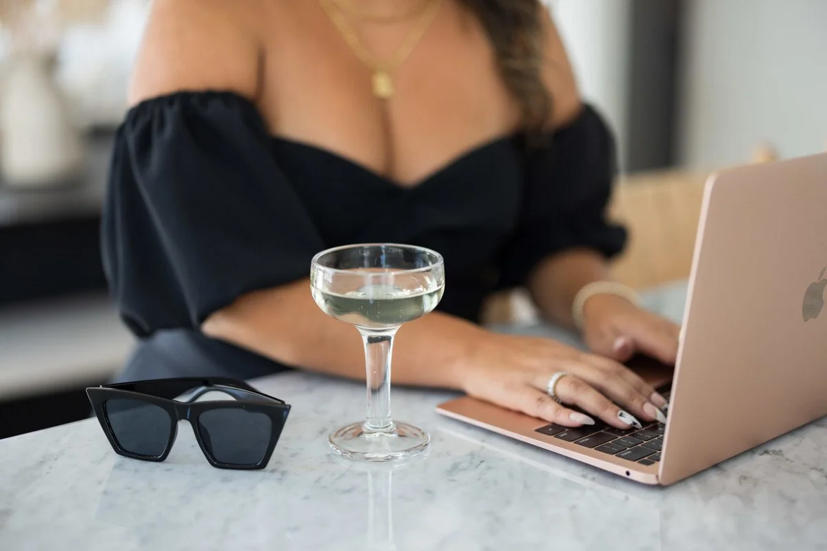 Close-up of a woman typing on a rose-gold laptop with a glass of champagne and black sunglasses on a marble table — chic lifestyle imagery for an online business coach working from home.