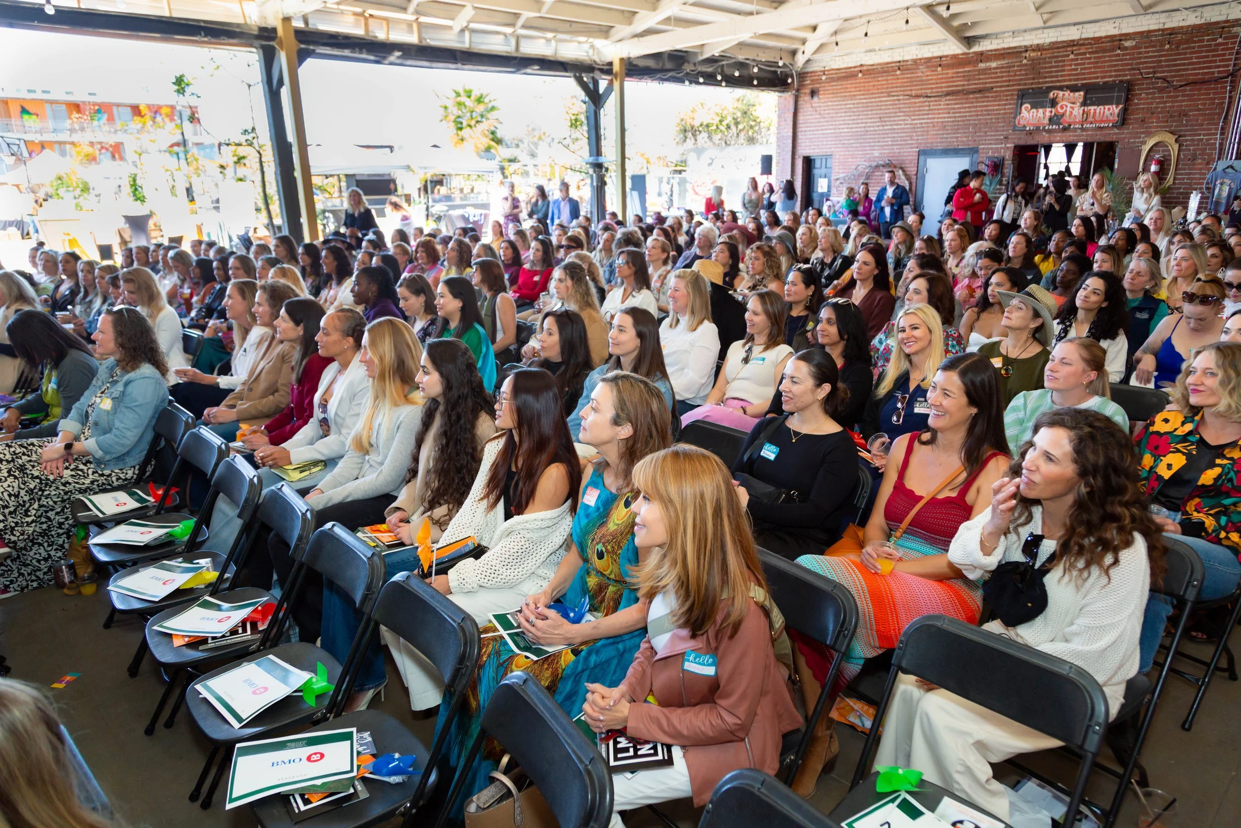 Large group of women gathered in a lively event, sitting attentively in a bright, open space. The crowd is diverse, with women engaged in the presentation or discussion, reflecting a sense of community, empowerment, and collaboration.