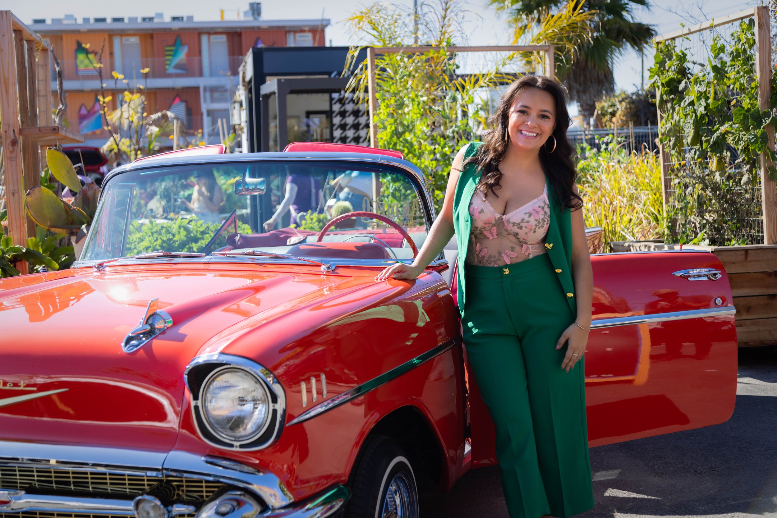 Jazmin smiling confidently next to a vintage red car, wearing a green vest and pants with a floral top. The background features vibrant greenery and modern urban elements, reflecting a stylish and energetic vibe.