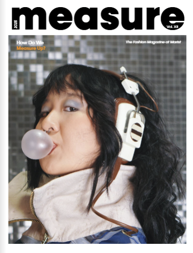 A woman with black curly hair blowing a bubblegum bubble, wearing vintage headphones and a beige high-collared jacket, standing in front of a tiled wall.