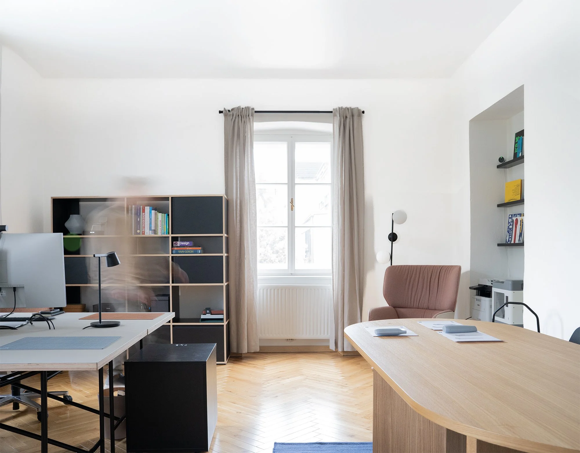 Ein modernes Büro mit Holzfußboden, weißen Wänden und vielen Fenstern. Es gibt Tische mit Computern, Regale mit Büchern und Dekorationsgegenständen, eine Stehlampe, und bequeme Stühle.
