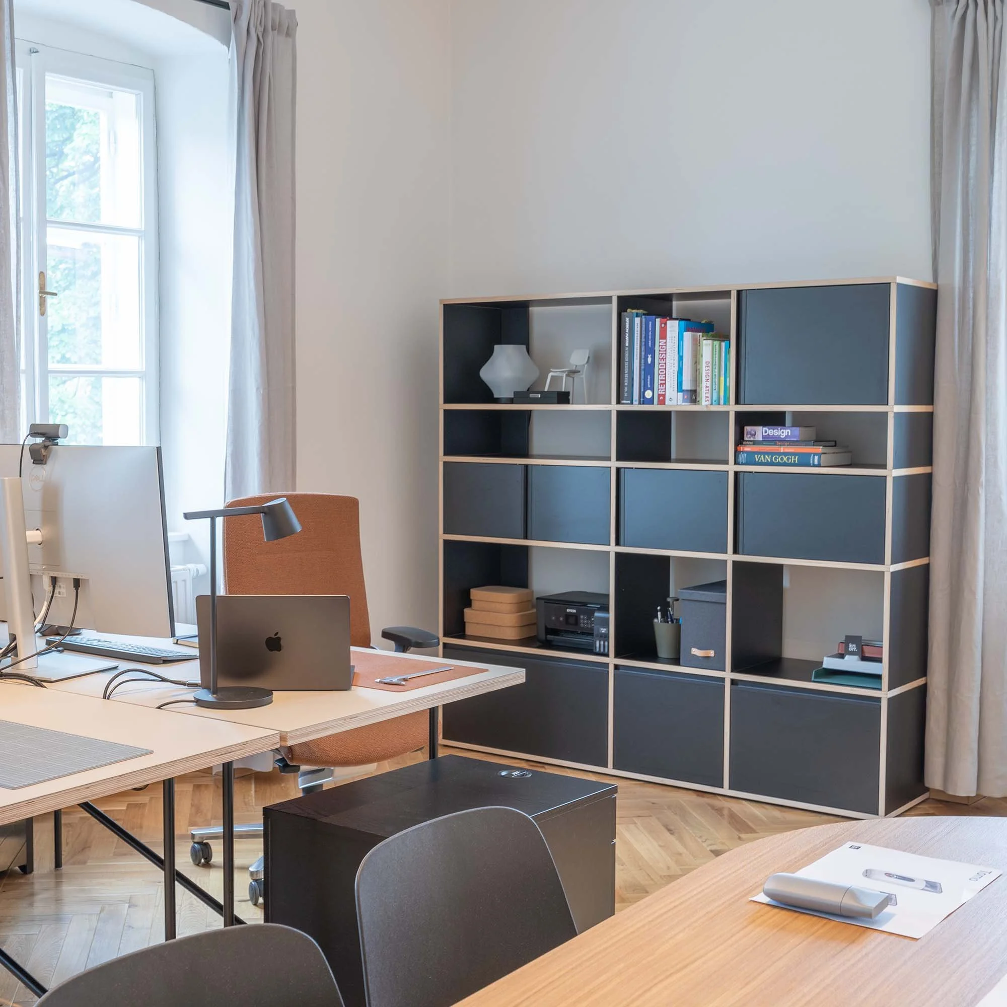 Ein Büro mit einem großen Fenster, weißen Wänden und einem schwarzen Regalschrank, der Bücher, eine Lampe und andere Gegenstände enthält. Es gibt einen Holztisch mit mehreren Stühlen und einen Computer samt Zubehör.