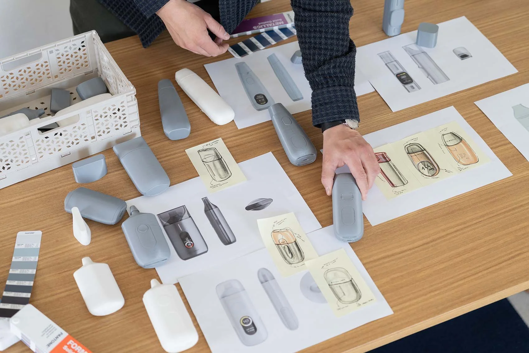 Ein Tisch mit mehreren skizzen und Produktmodellen von elektrischen Geräten sowie Farbfächern. Es werden Menschen bei der Gestaltung und Entwicklung eines Produkts beobachtet.