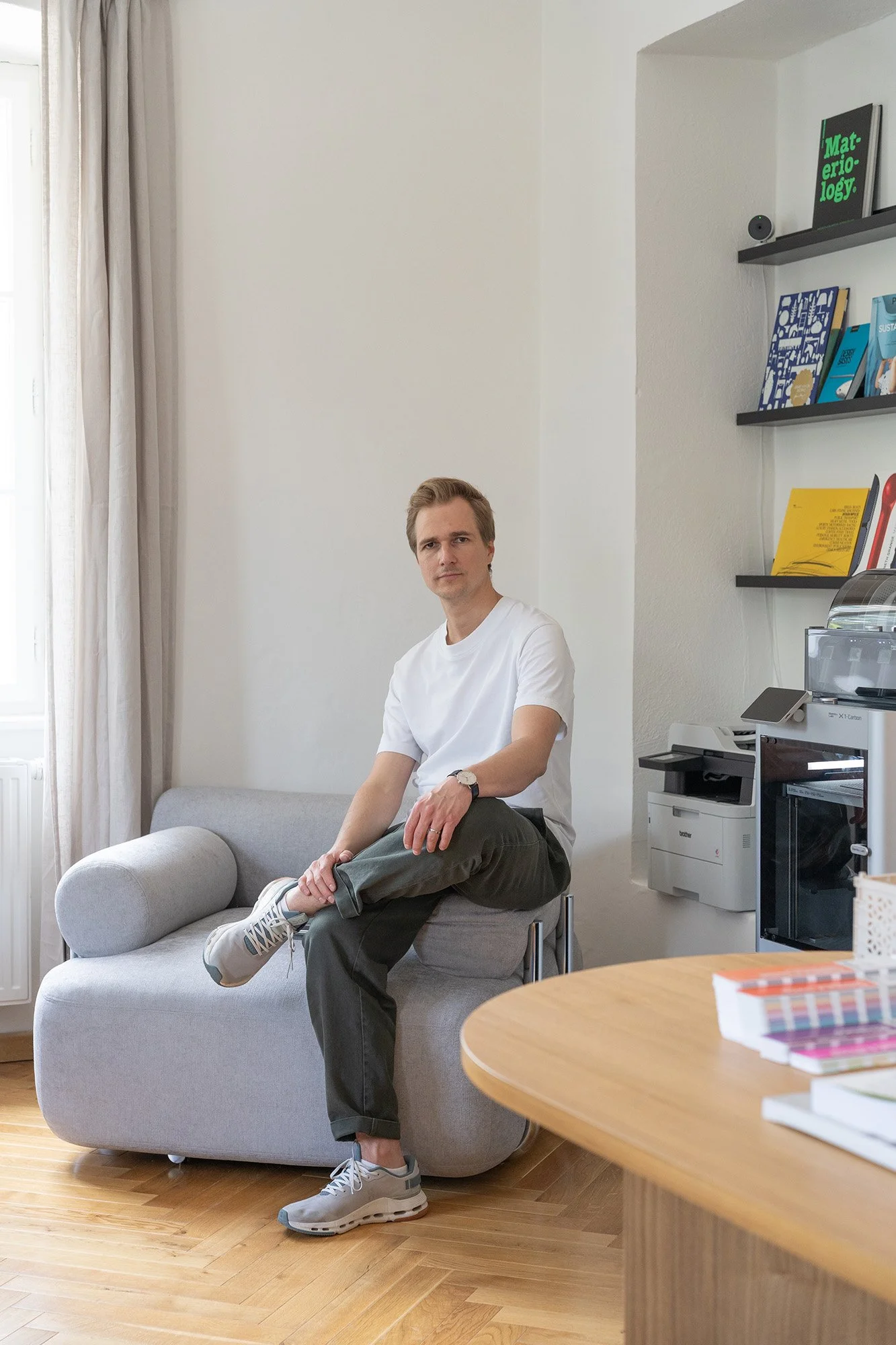 Junge Frau sitzt entspannt auf der Couch in einem modernen, hellen Raum mit Büchern und Regalen an der Wand, neben einem runden Tisch mit bunten Büchern.