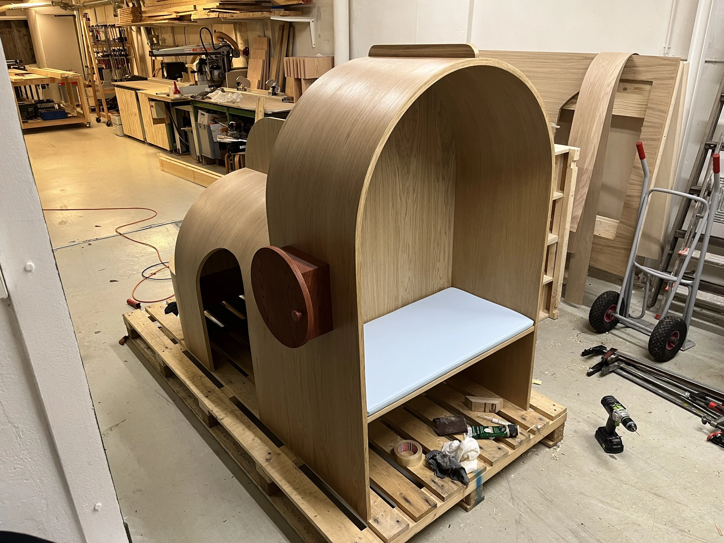 Udsnit af et værksted med træmøbler under konstruktion, særligt en træhylde eller et møbel med buede elementer i fokus, omgivet af værktøj og træmaterialer.