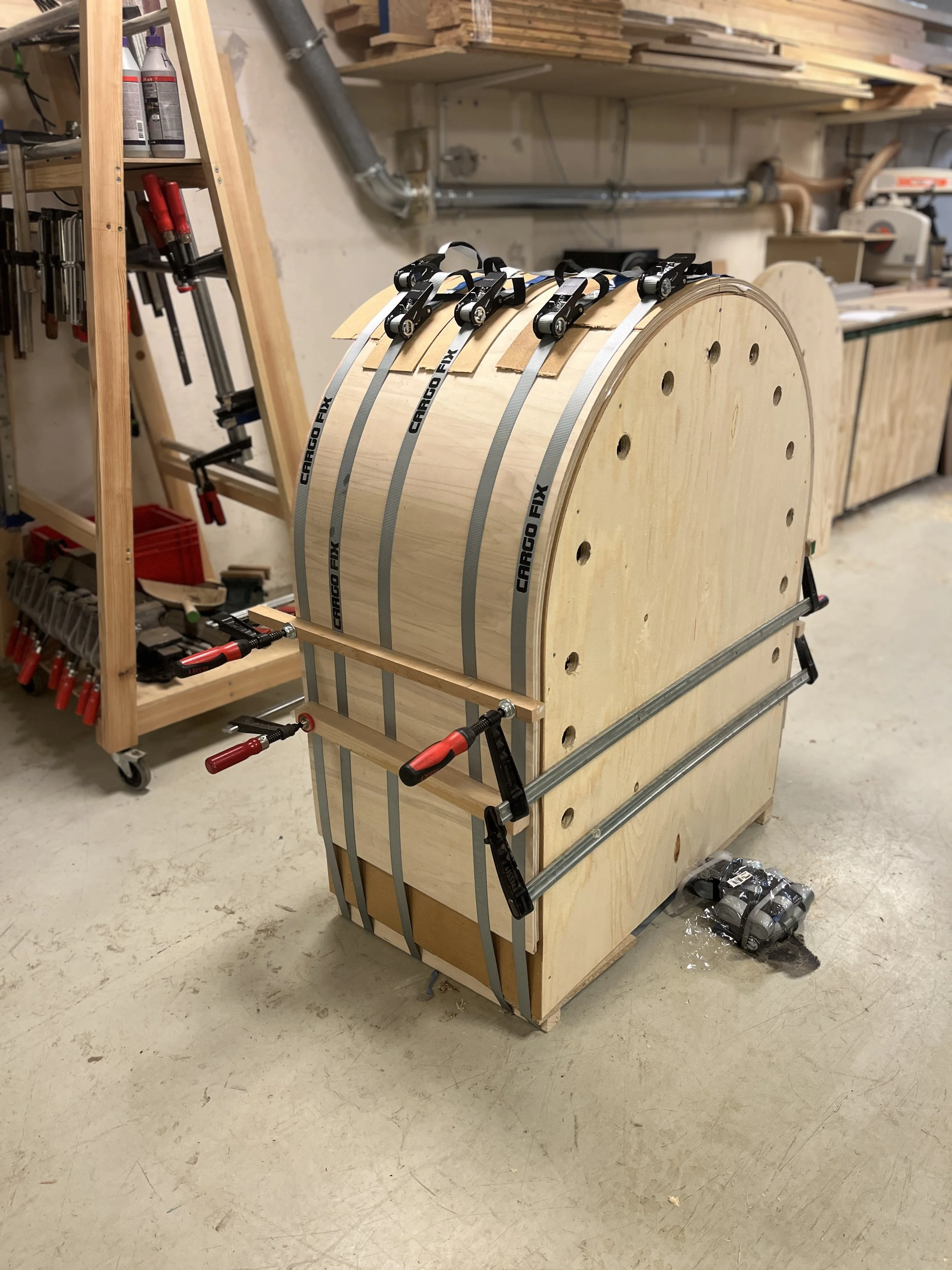 Et værksted med træmaterialer og værktøj, der er i gang med at bygge en stor træskive, der er spændt fast med metalbånd, sandsynligvis en del af en maskine eller møbel.