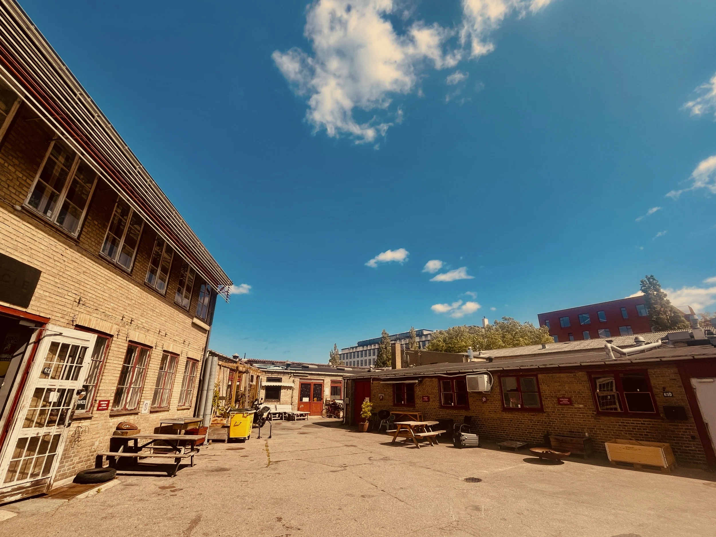 Et gårdmiljø med gamle murstensbygninger, borde, stole, og forskellige affalds- og cykeludstyr, under en klar blå himmel med enkelte skyer.
