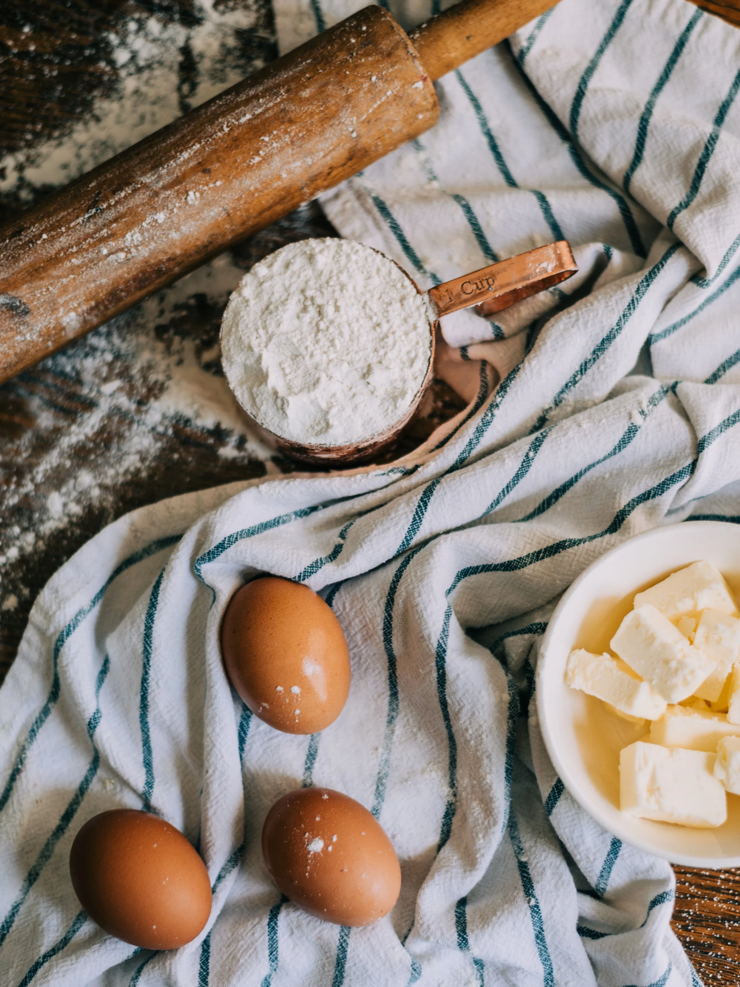 Kitchen scene with flour in a measuring cup, eggs on a striped cloth, a rolling pin, and butter in a bowl.