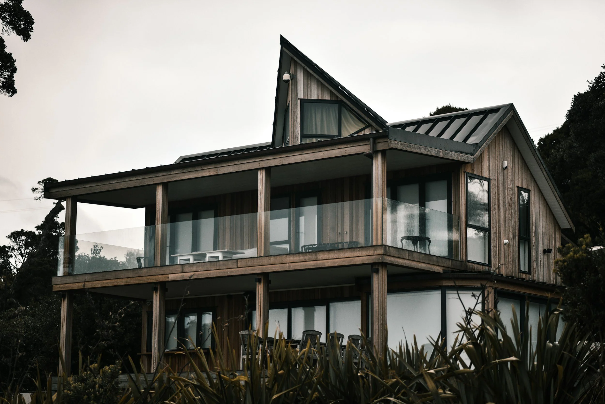 Maison moderne en bois à plusieurs niveaux, avec grandes fenêtres et balcon en verre, entourée de végétation, sous un ciel nuageux.