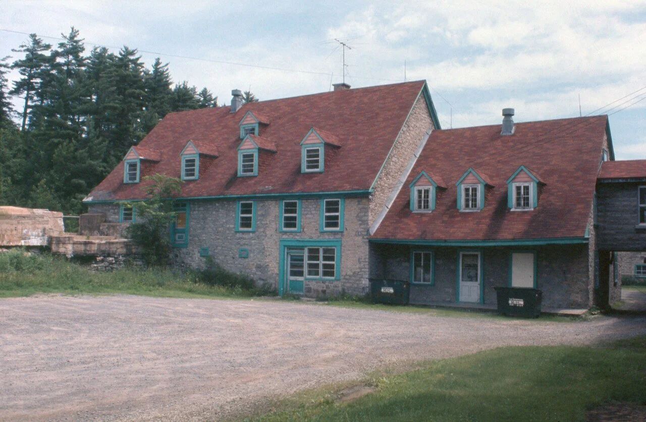 Une maison en pierre avec un toit en tuiles rouges et plusieurs fenêtres. La maison est située dans un espace vert avec des arbres en arrière-plan.