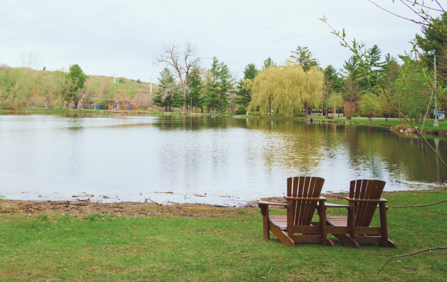 Deux chaises en bois sur l'herbe au bord d'un lac avec arbres et un parc au fond.