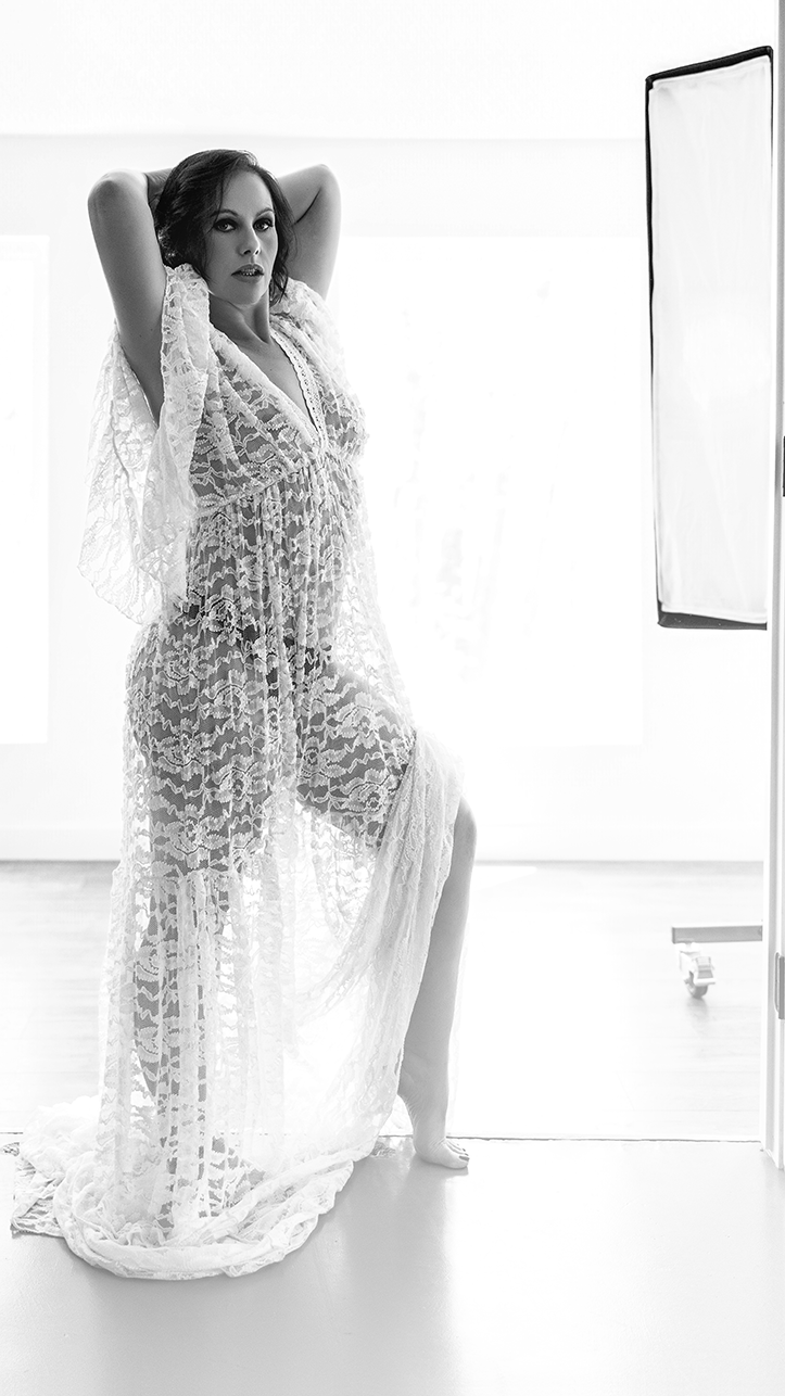 A woman standing in front of a window wearing a long, sheer lace dress, with one hand behind her head and the other resting on her hip, in front of a studio light.