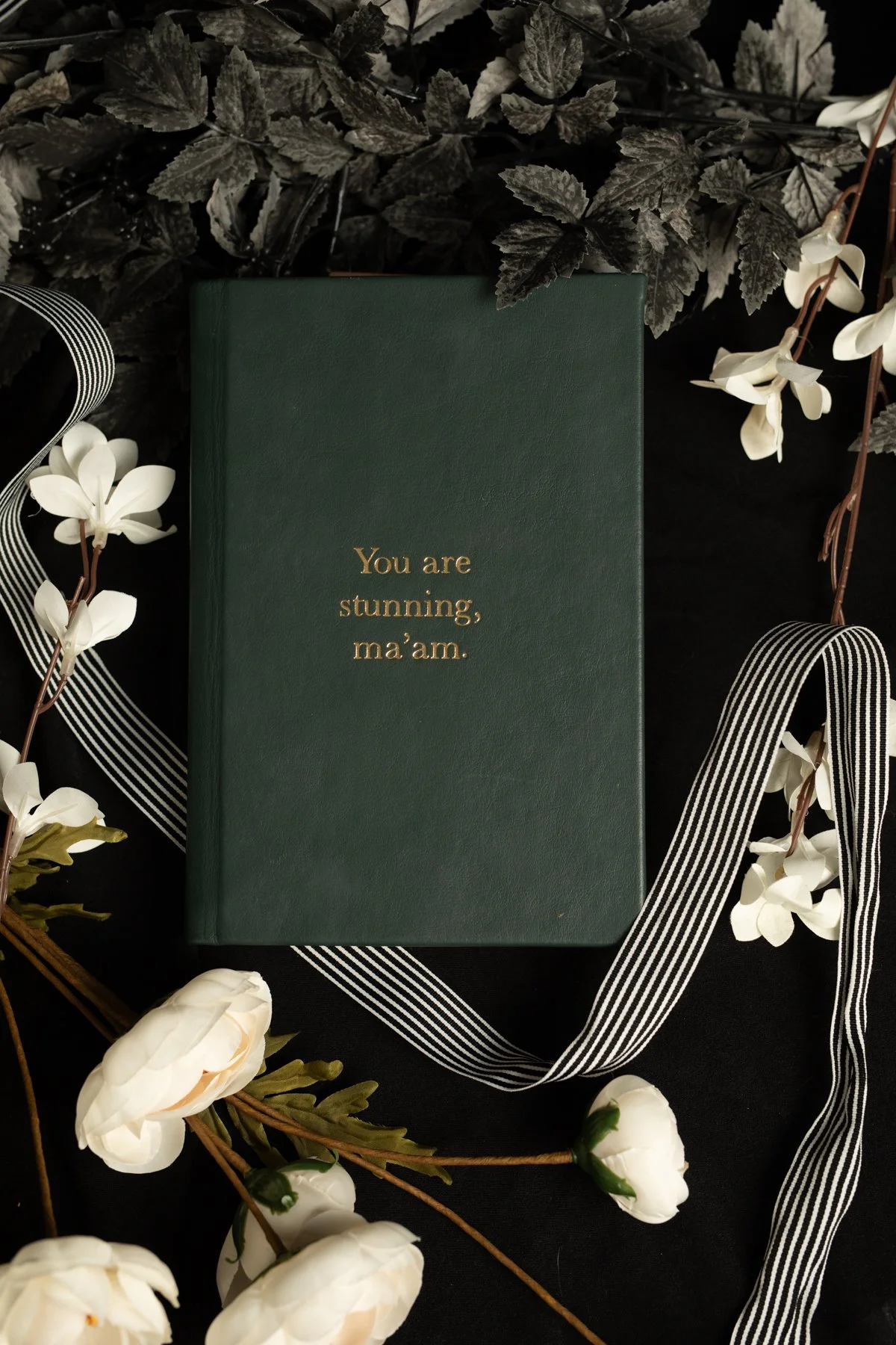 A dark green book with the gold embossed words "You are stunning, ma’am" on the cover, surrounded by white flowers, black and white striped ribbon, and black leaves, set against a black background.