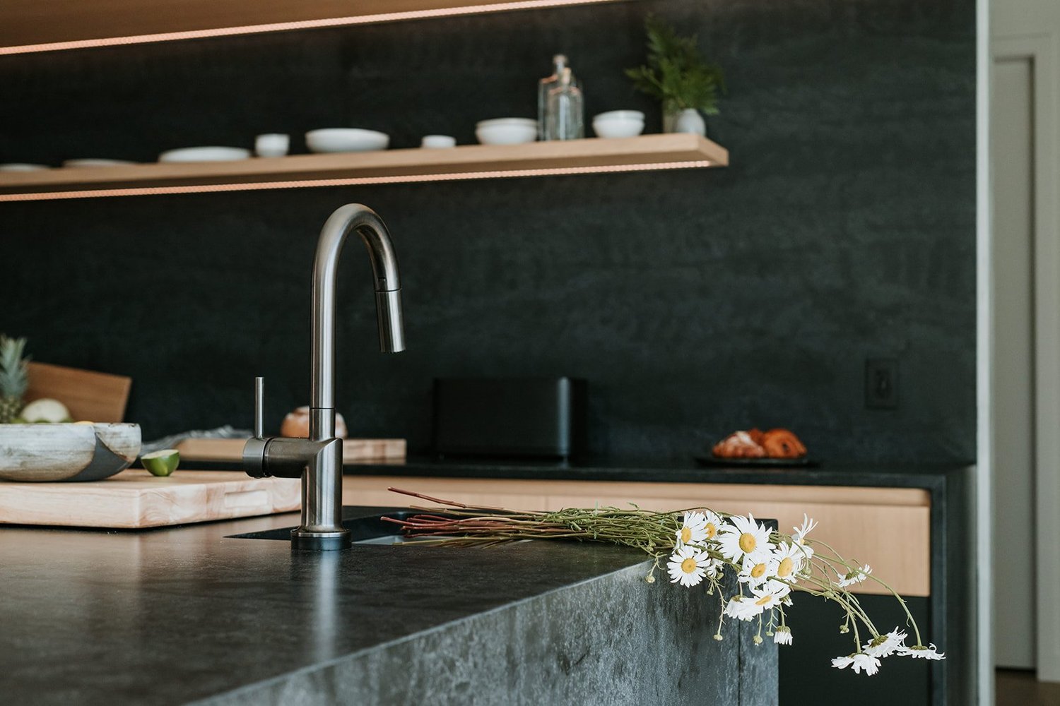 Sunstone Kitchen faucet close-up on dark stone, daisies, exposed shelf behind — new-build interior design in Pemberton, BC by LRD Studio. Photography by Darby Magill.