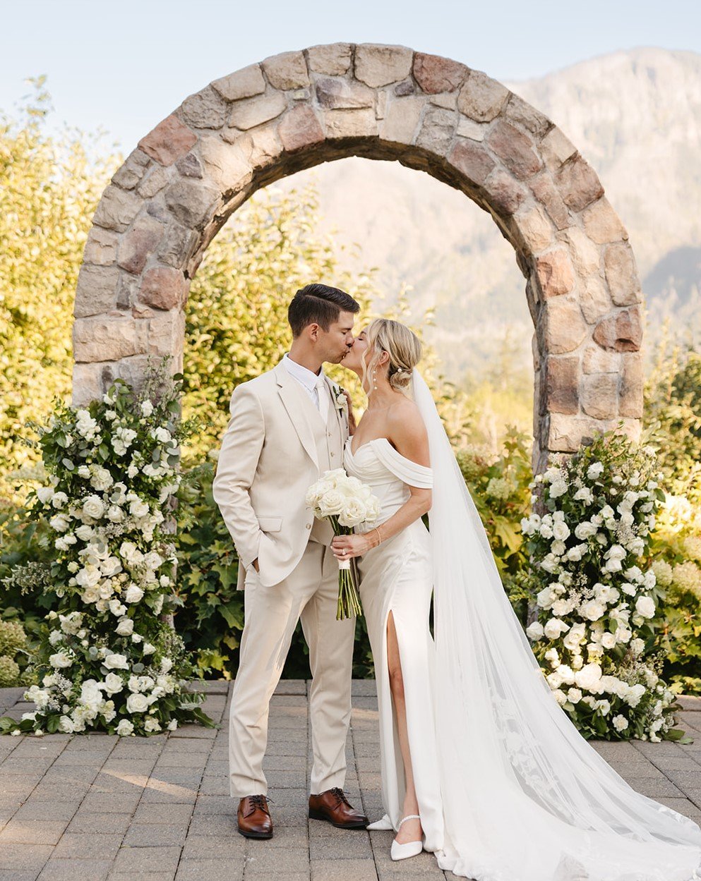 A bride and groom kiss under a stone arch during their outdoor wedding, surrounded by white floral arrangements and greenery with mountain scenery in the background.