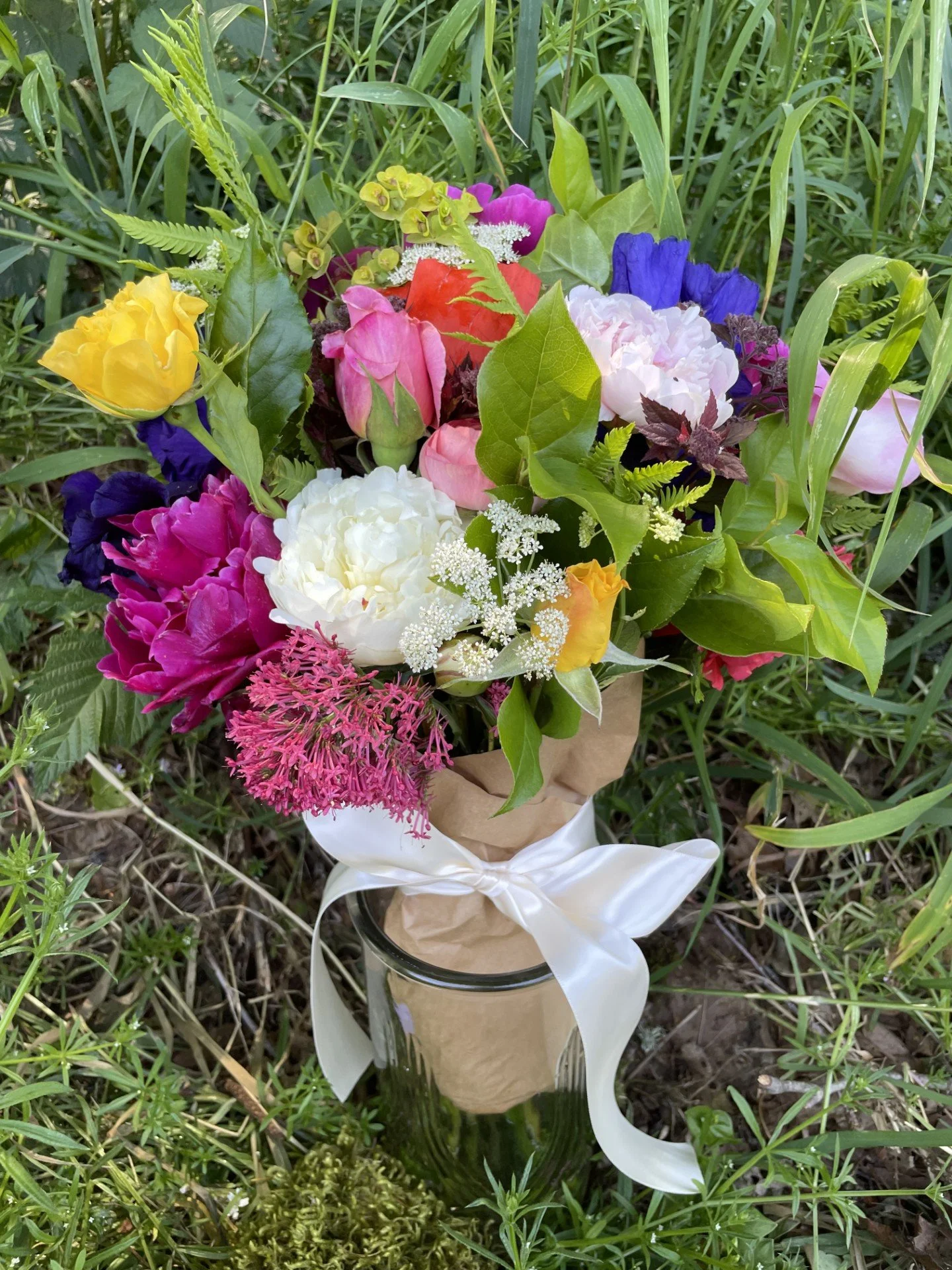 A colorful bouquet of various fresh flowers wrapped in brown paper and tied with a white ribbon, placed in a glass vase surrounded by green grass and foliage.