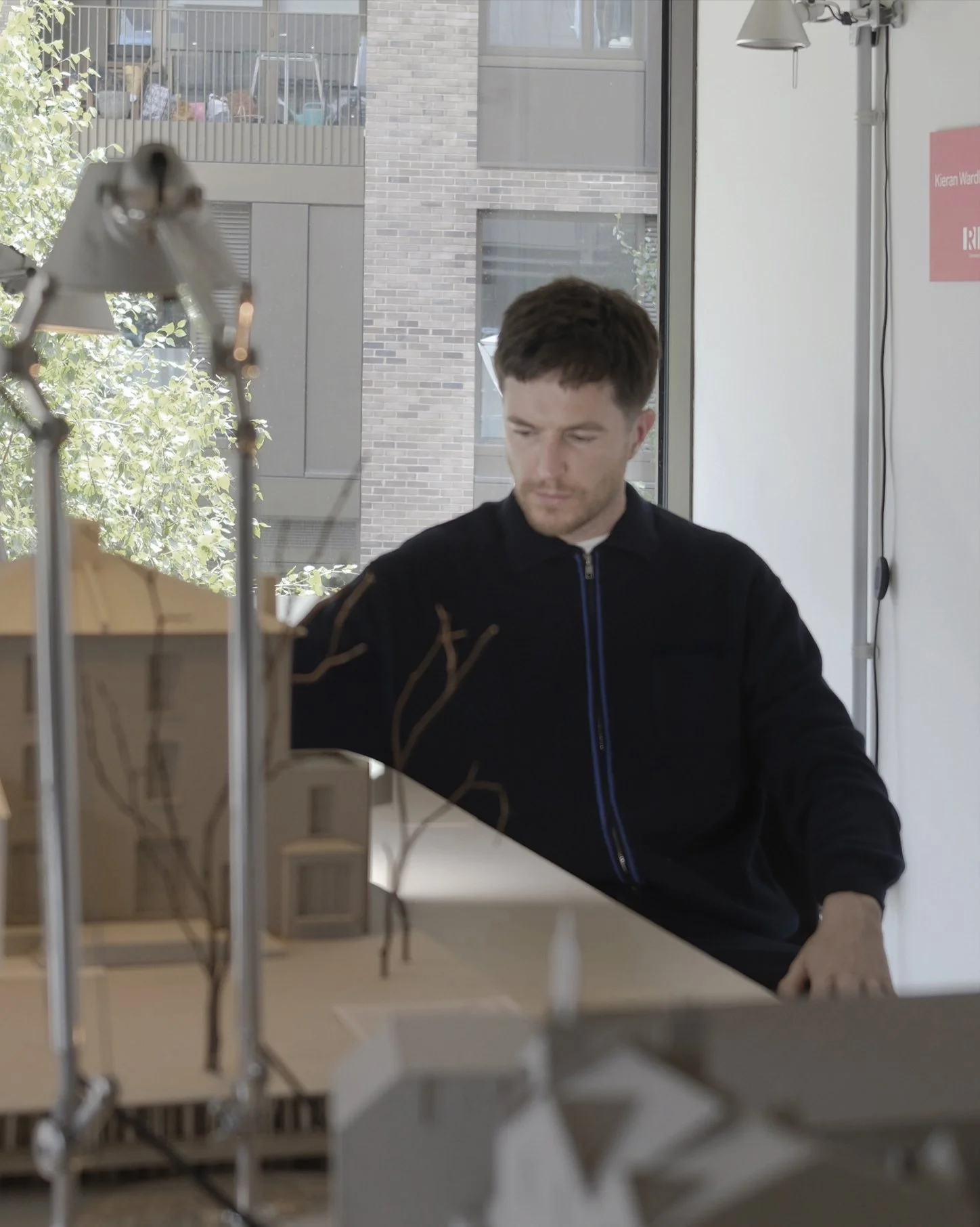 A man with short brown hair and a beard, wearing a black jacket with a blue zipper, is working on a project at a table indoors near a large window with a view of a brick building and trees outside.