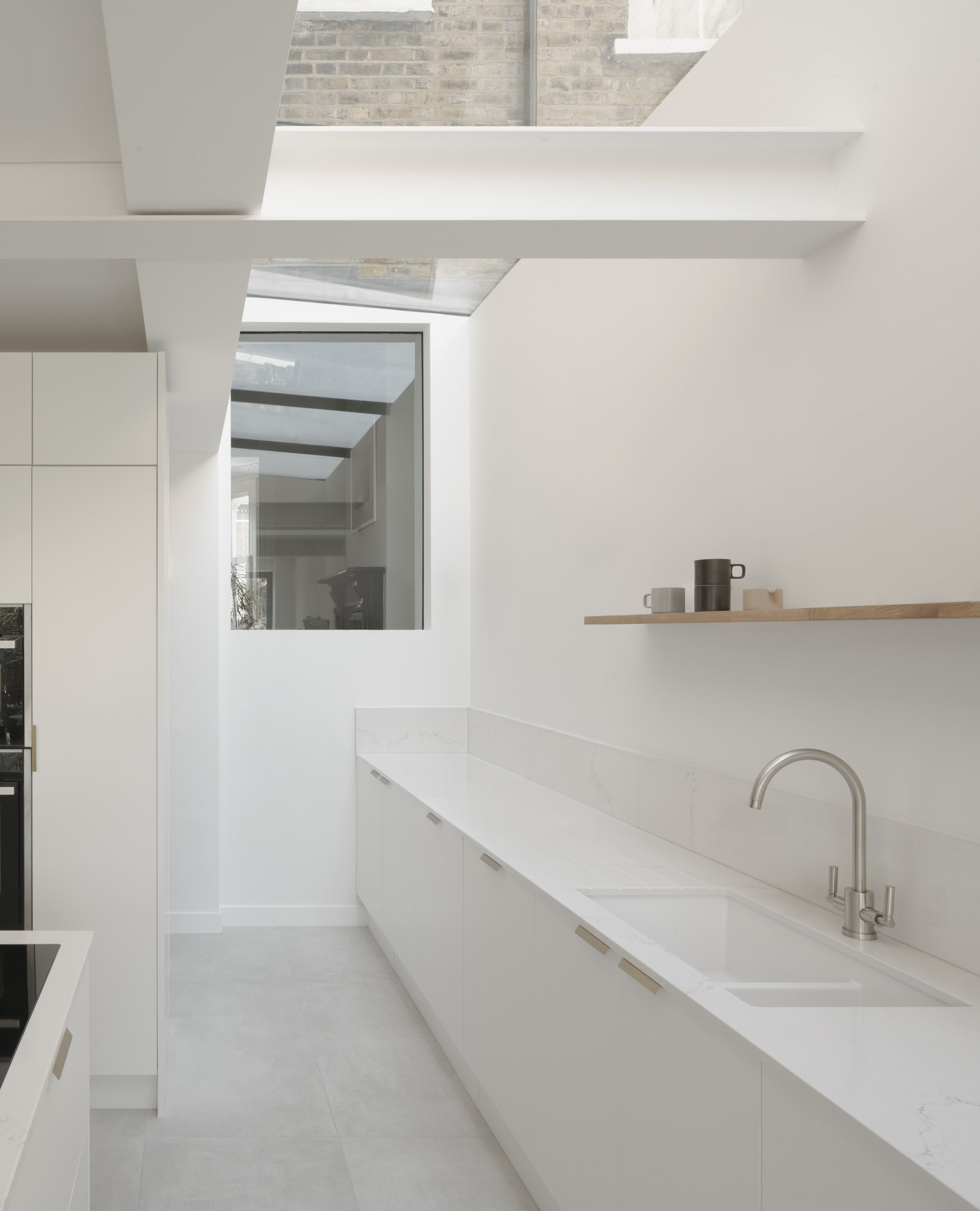 Kieran Wardle Architects — Modern minimalist white kitchen with a marble countertop, stainless steel faucet, and a floating wooden shelf with cups, large window, and exposed bri