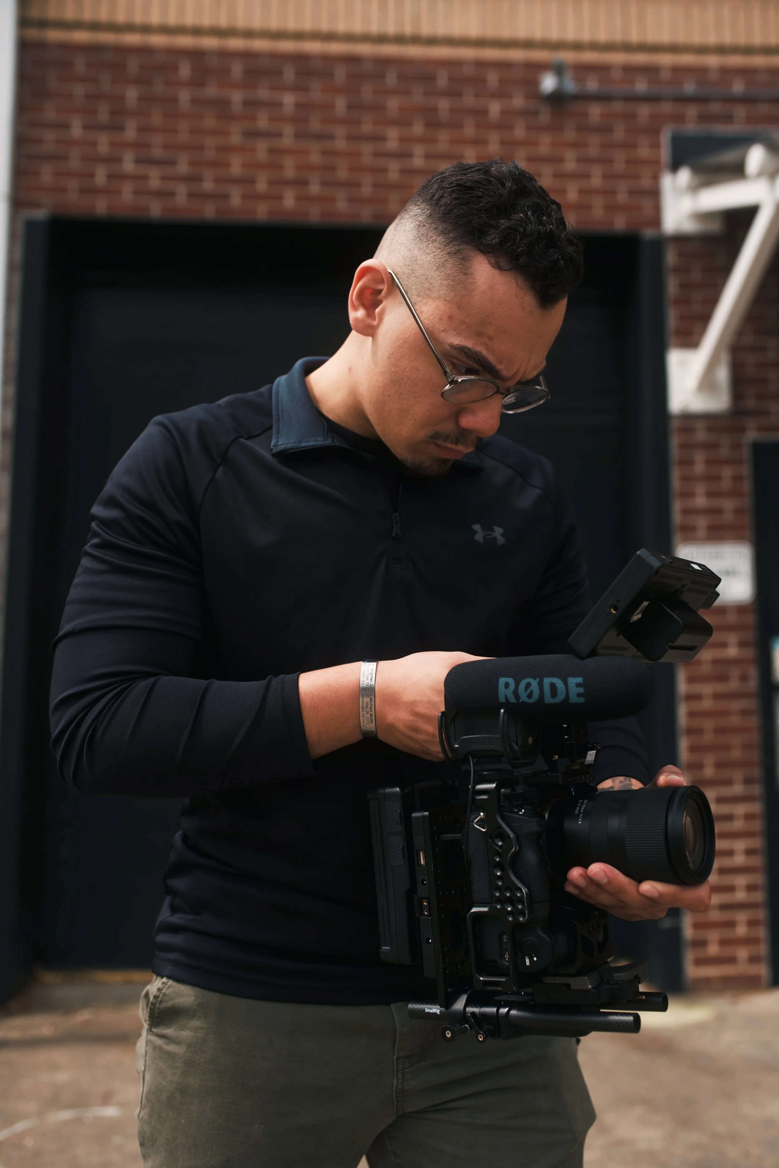 A man wearing glasses and a black athletic hoodie operating a professional video camera with a Rode microphone attached, standing in front of a brick building.