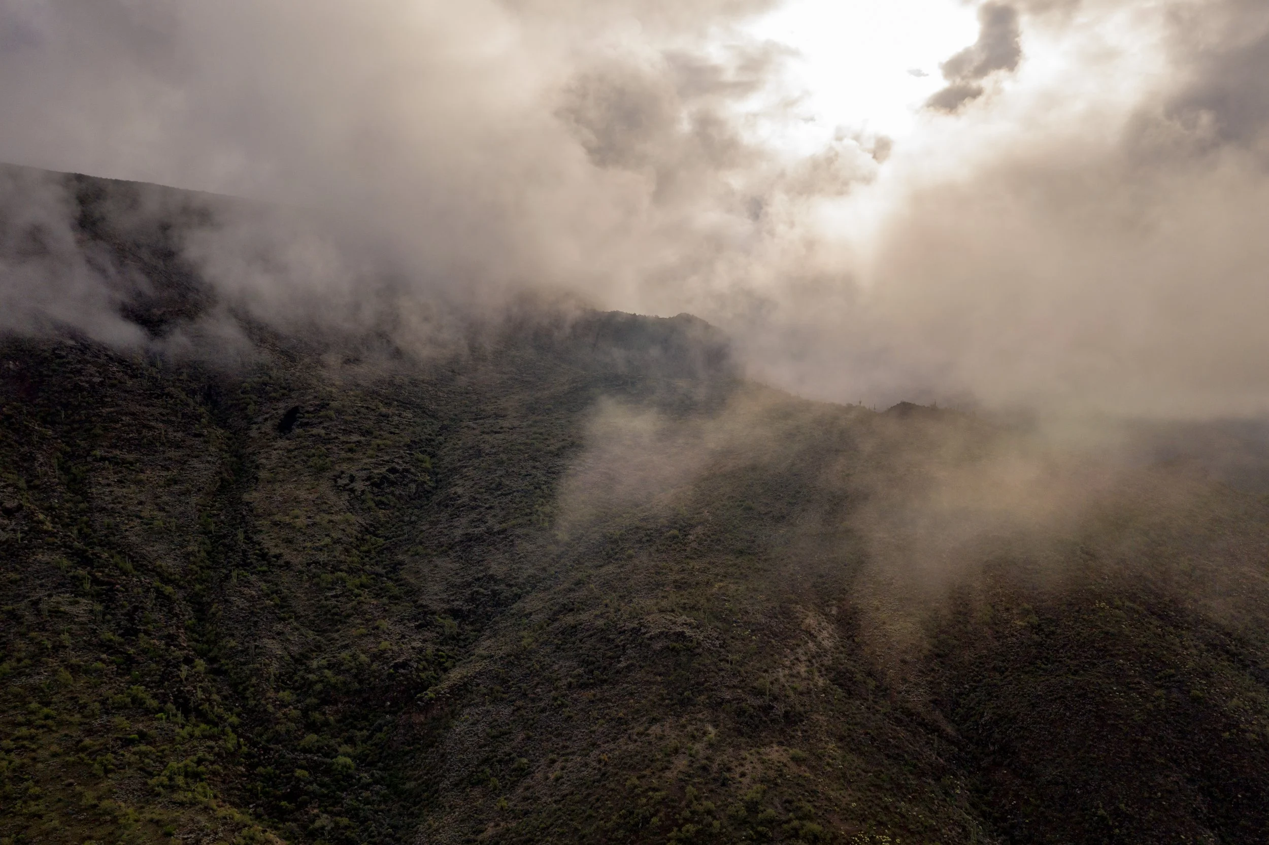 A mountain landscape covered in fog and clouds with a partly cloudy sky and sunlight breaking through.