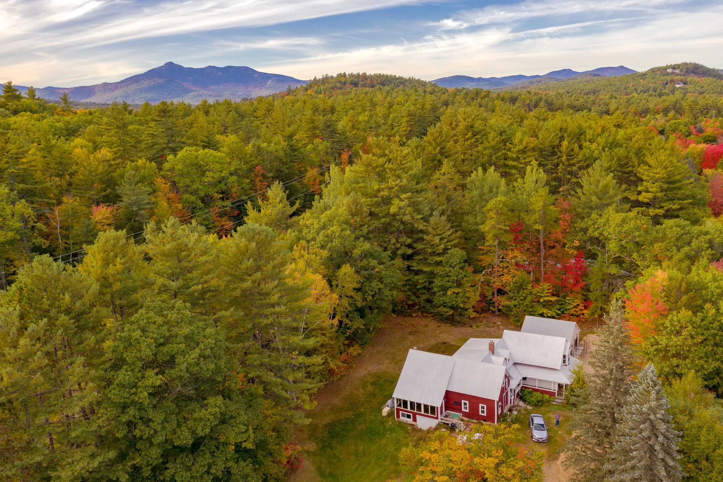 Aerial view of a rural landscape with colorful fall foliage, a white house, and distant mountains under a partly cloudy sky.