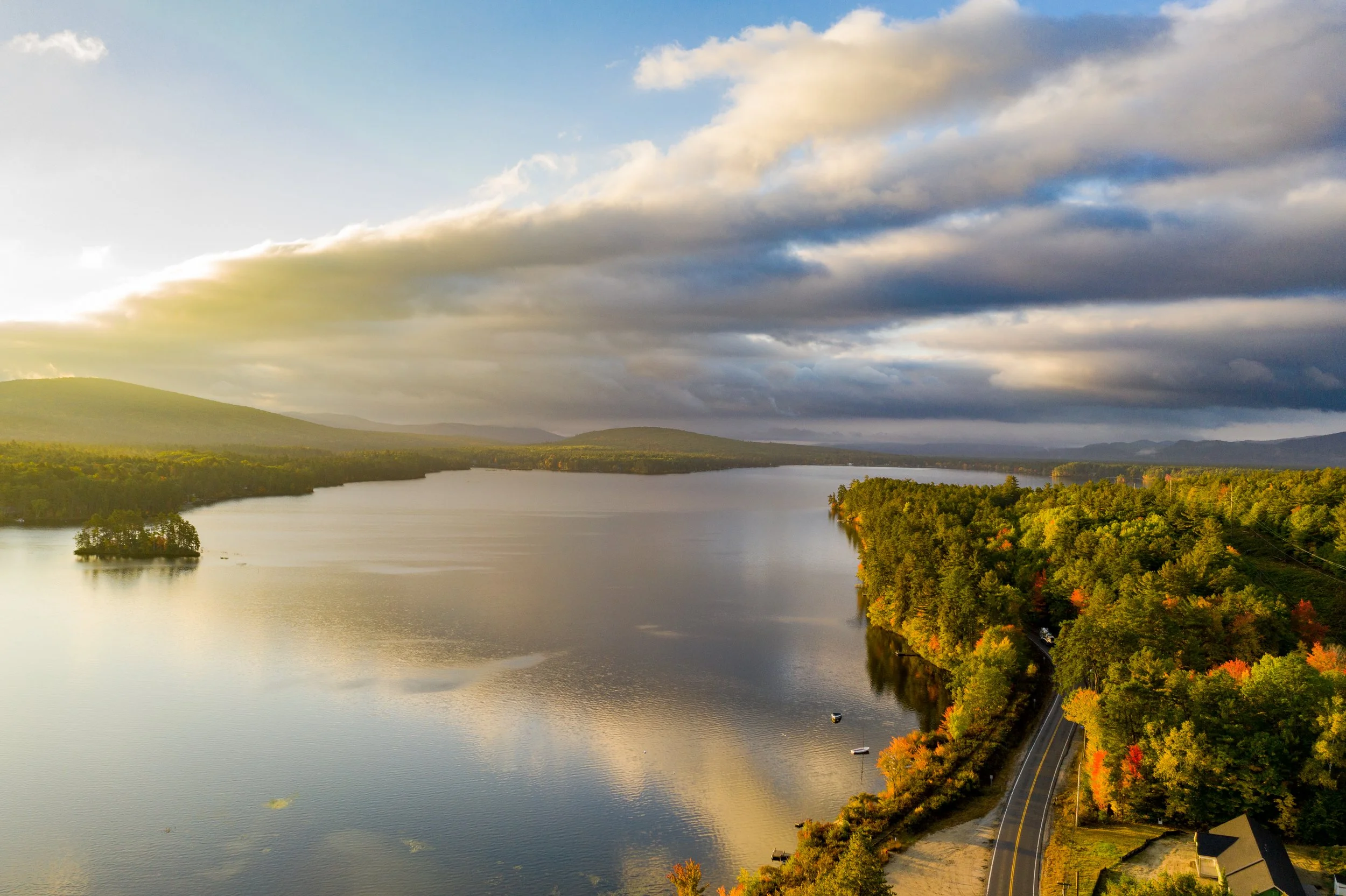 A large river with calm water reflecting the sky, surrounded by green and autumn-colored trees, hills in the background, and a cloudy sky overhead.