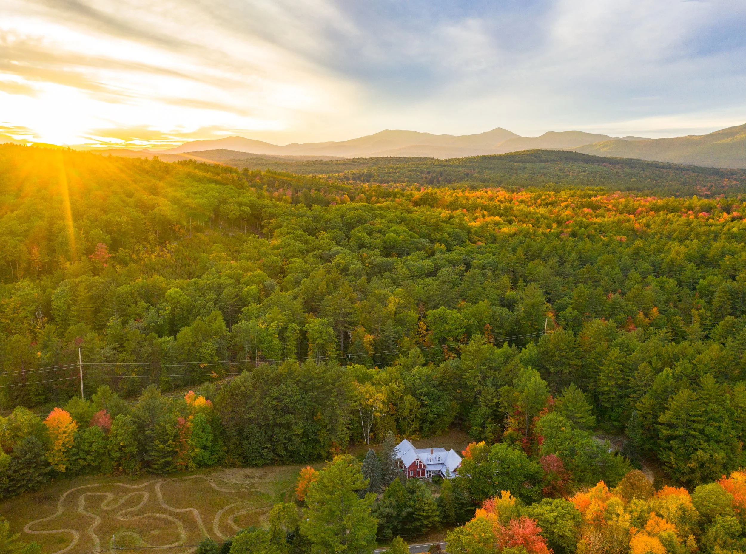 An aerial view of a rural landscape during sunset, featuring a dense forest with trees in fall colors, a white house with a dark roof, and mountains in the background under a partly cloudy sky.