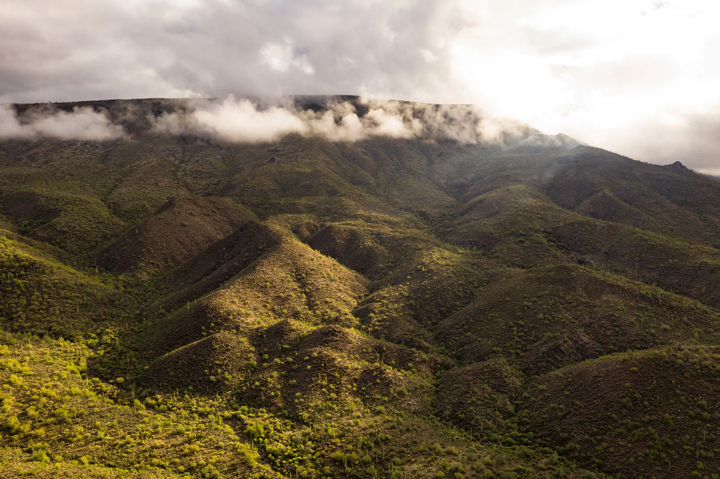 A mountainous landscape with green vegetation, partly covered by low hanging clouds and mist, under a cloudy sky.