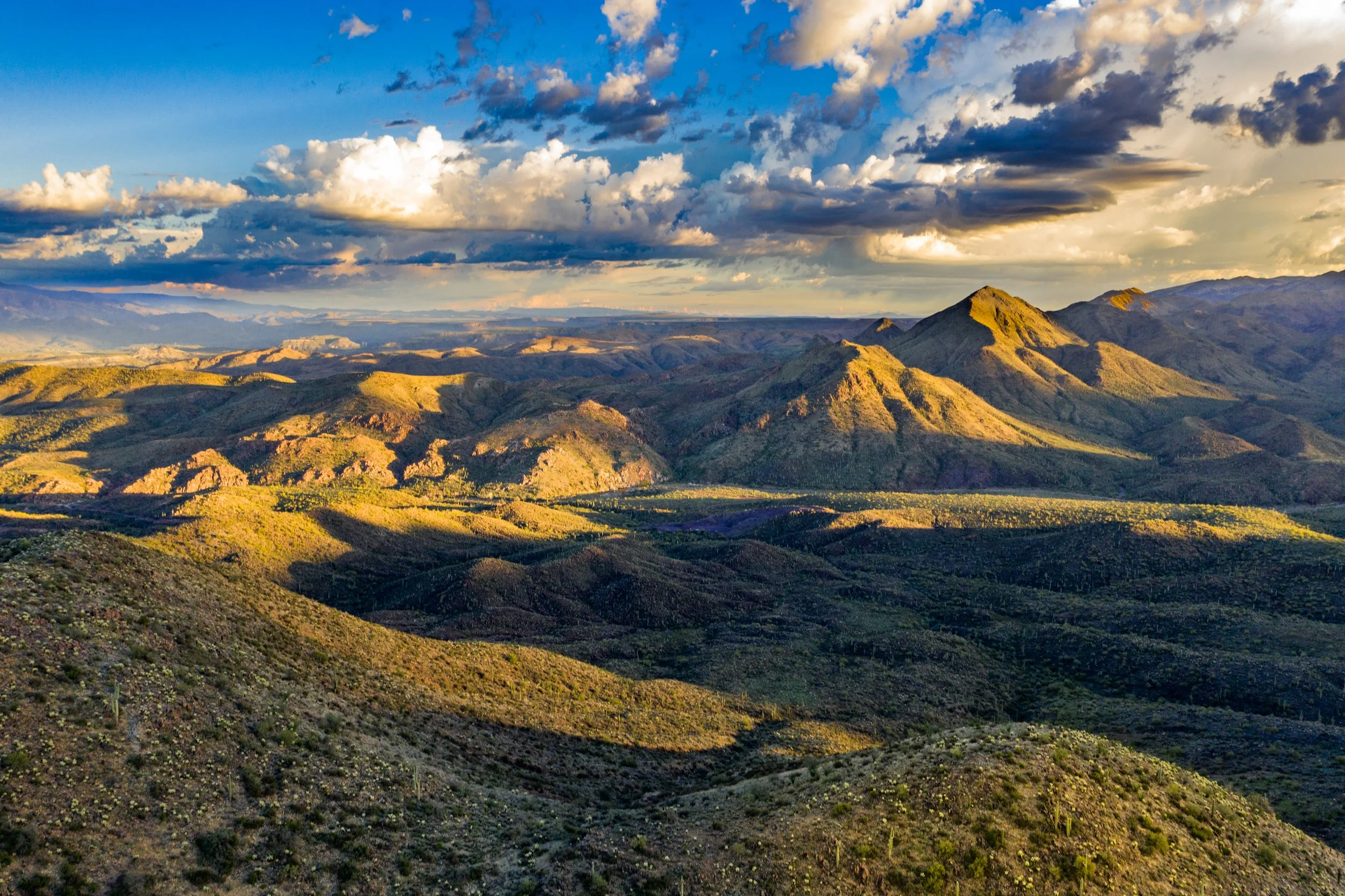 A landscape of rolling hills and mountains under a partly cloudy sky with sunlight illuminating parts of the terrain.