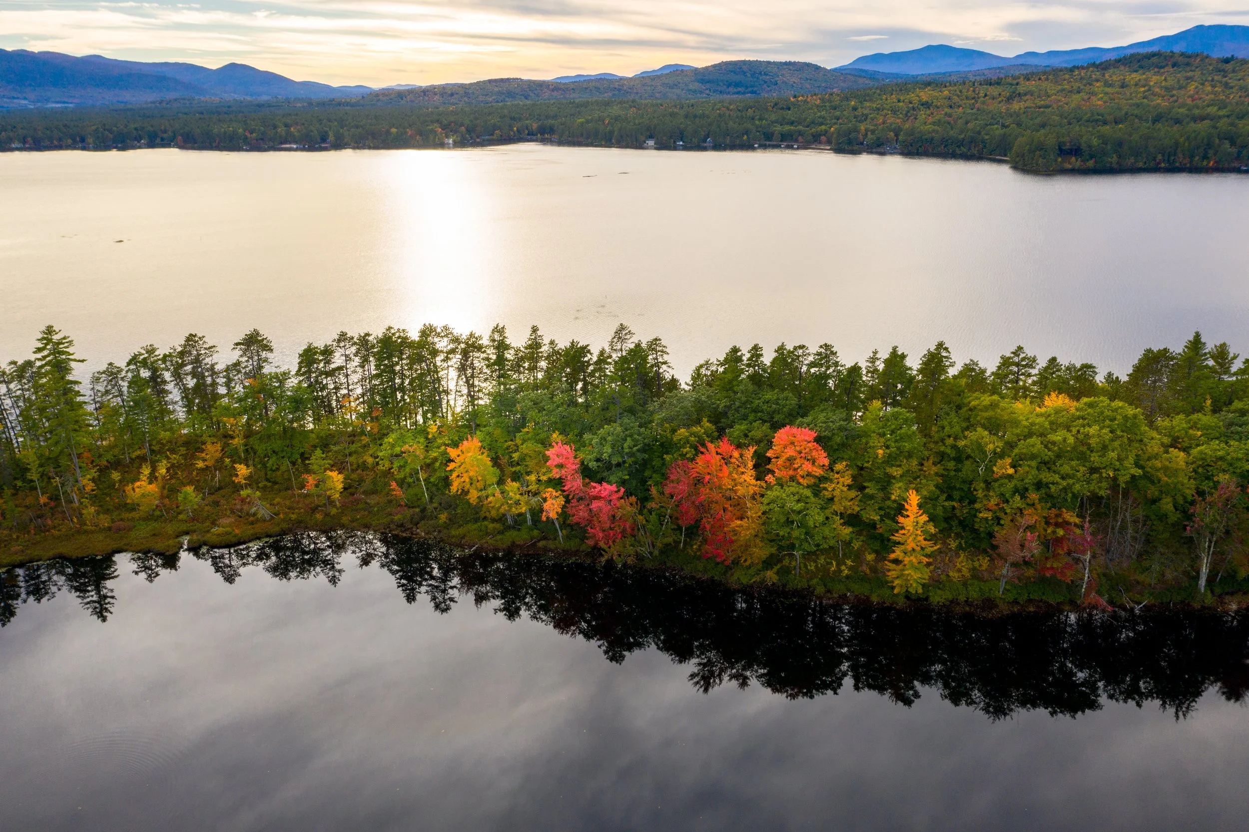 A scenic landscape featuring a large lake reflecting a cloudy sky, surrounded by a dense forest with trees showing fall colors like red, orange, and yellow, with mountains in the distance under a sunset or sunrise sky.