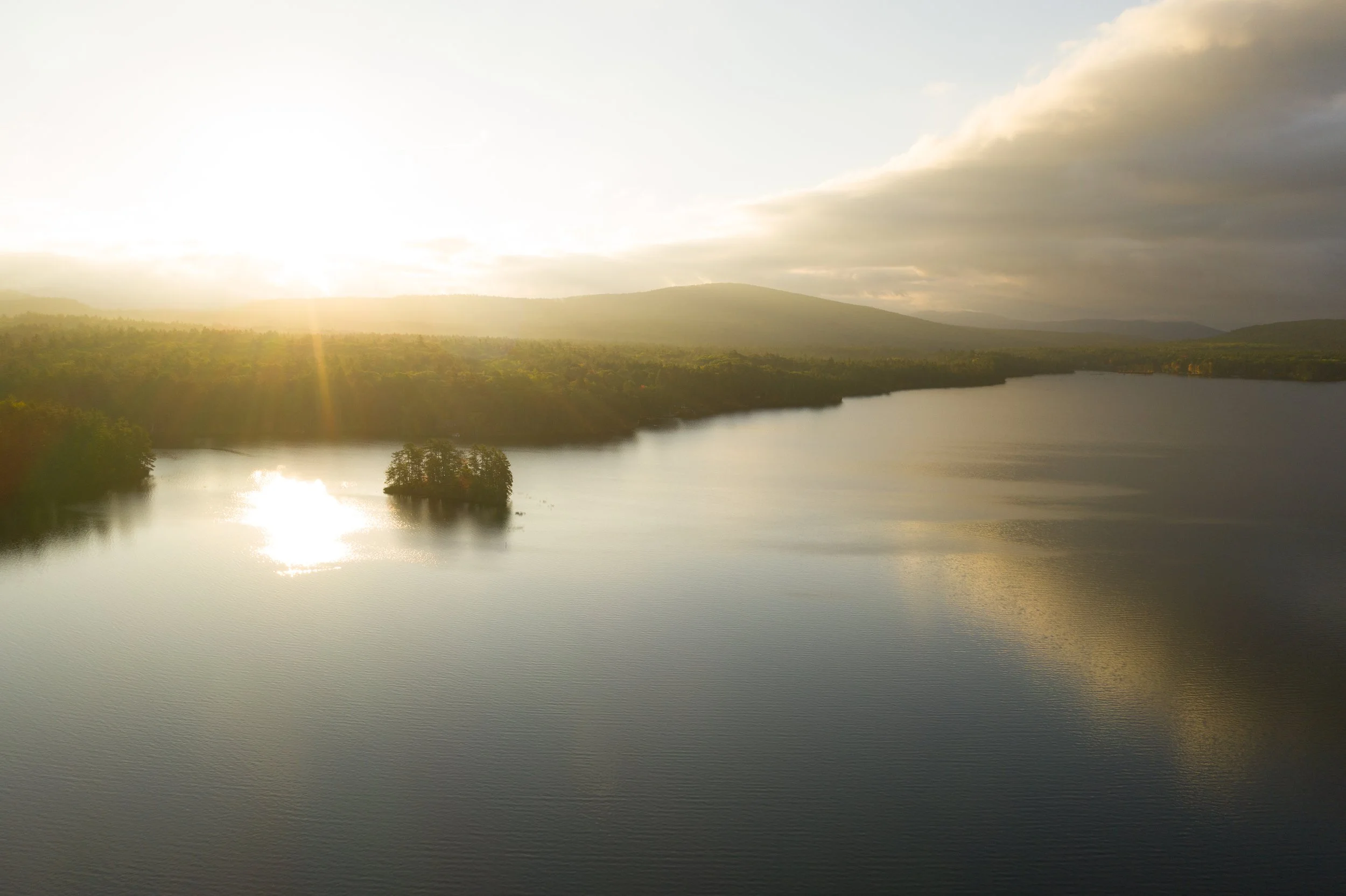 Sunset over a lake with a small island, surrounded by forested hills and partly cloudy sky.