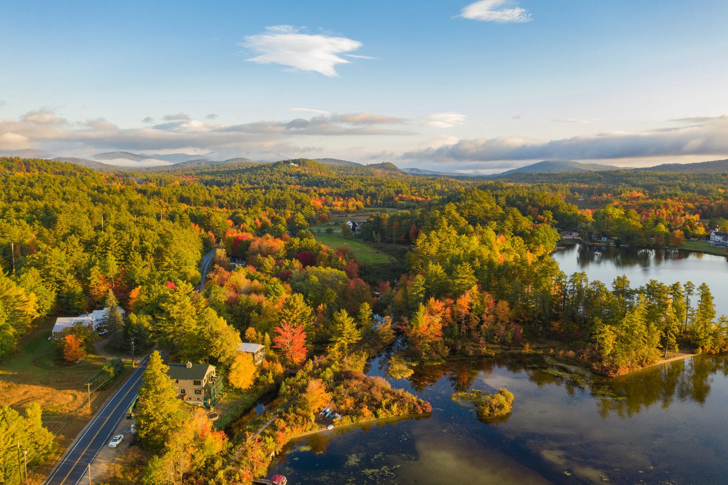 A scenic aerial view of a landscape with colorful autumn trees, a body of water, houses, and mountains in the distance under a partly cloudy sky.