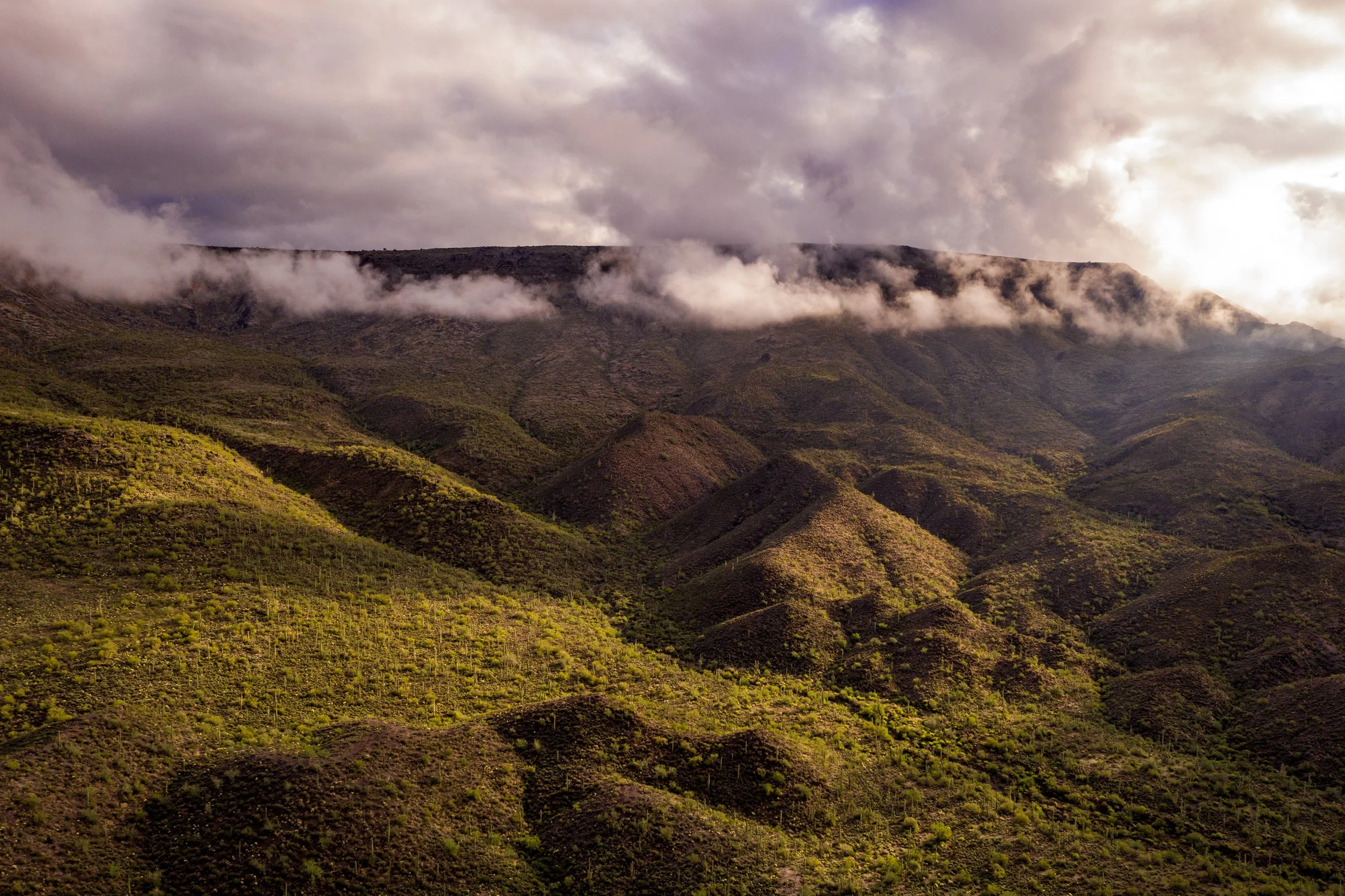 A mountainous landscape with green hills, low-hanging clouds or fog, and a cloudy sky.