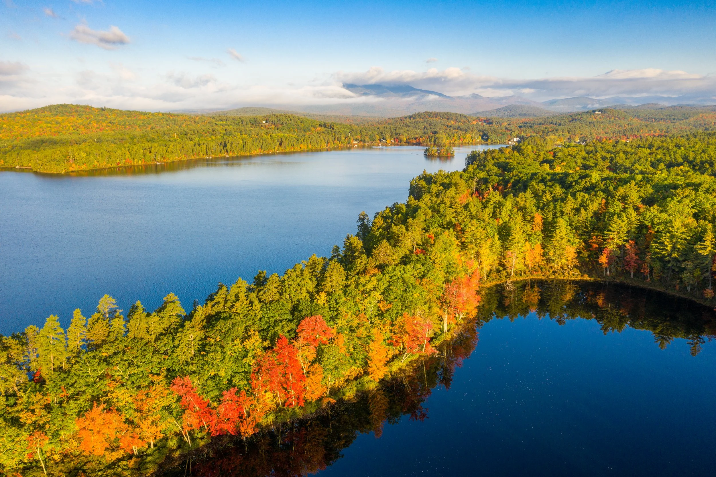 A scenic view of a lake surrounded by colorful autumn trees with mountains in the background and a clear blue sky.