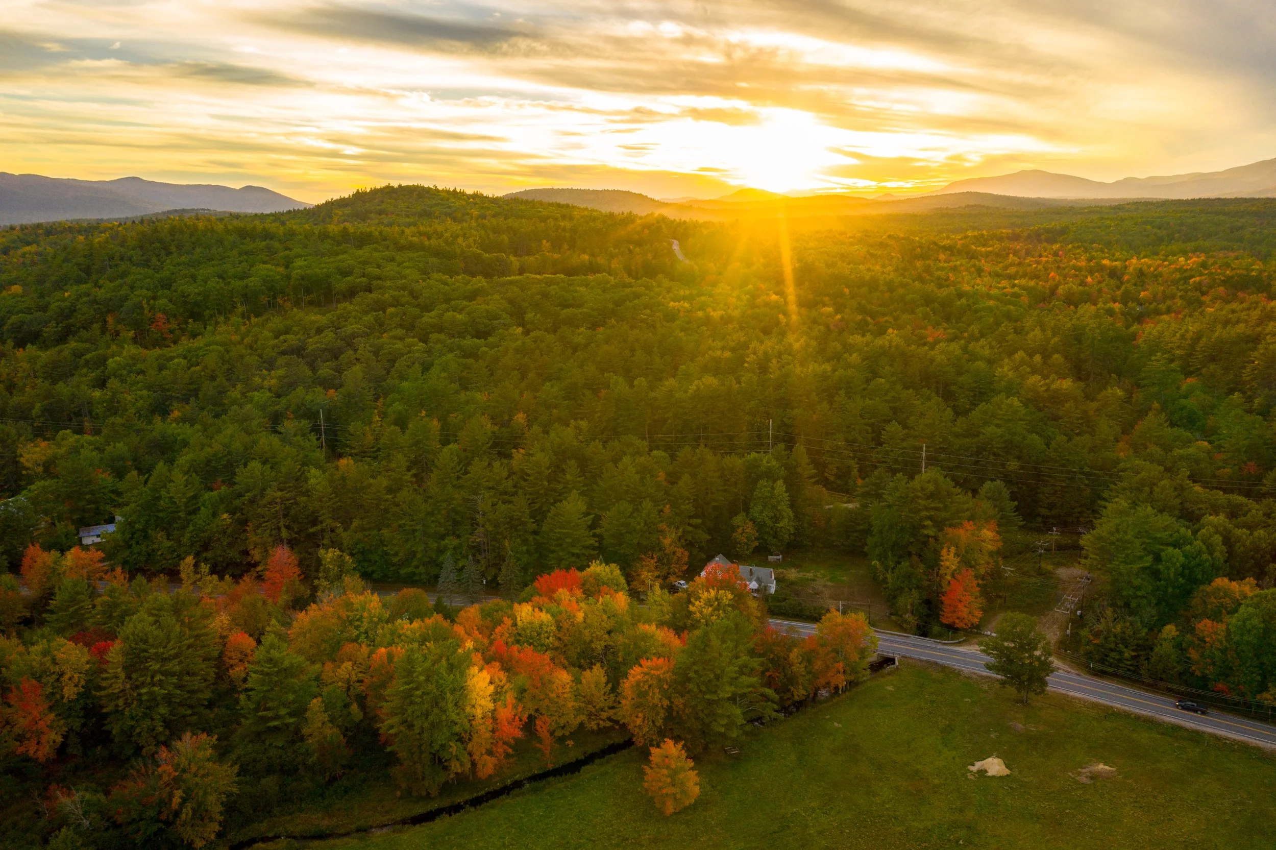 Sunset over a mountain landscape with dense green forest, a few houses, and a road in the foreground.