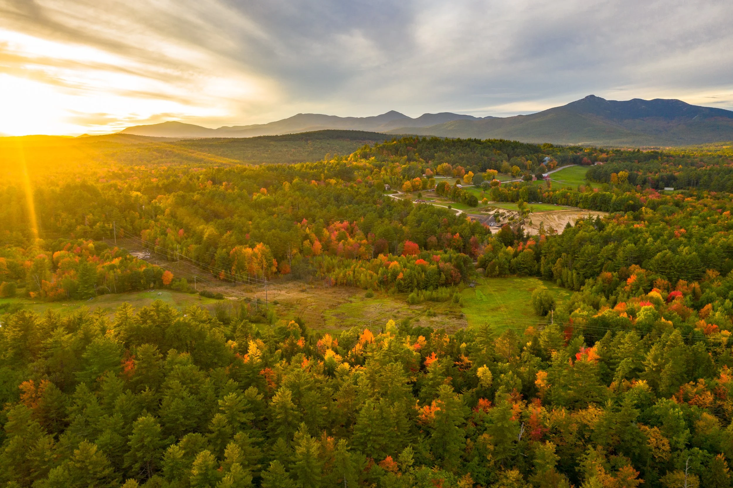 A scenic landscape of a forested valley with a mix of green and autumn-colored trees, rolling hills, and mountains in the distance under a partly cloudy sky during sunset.
