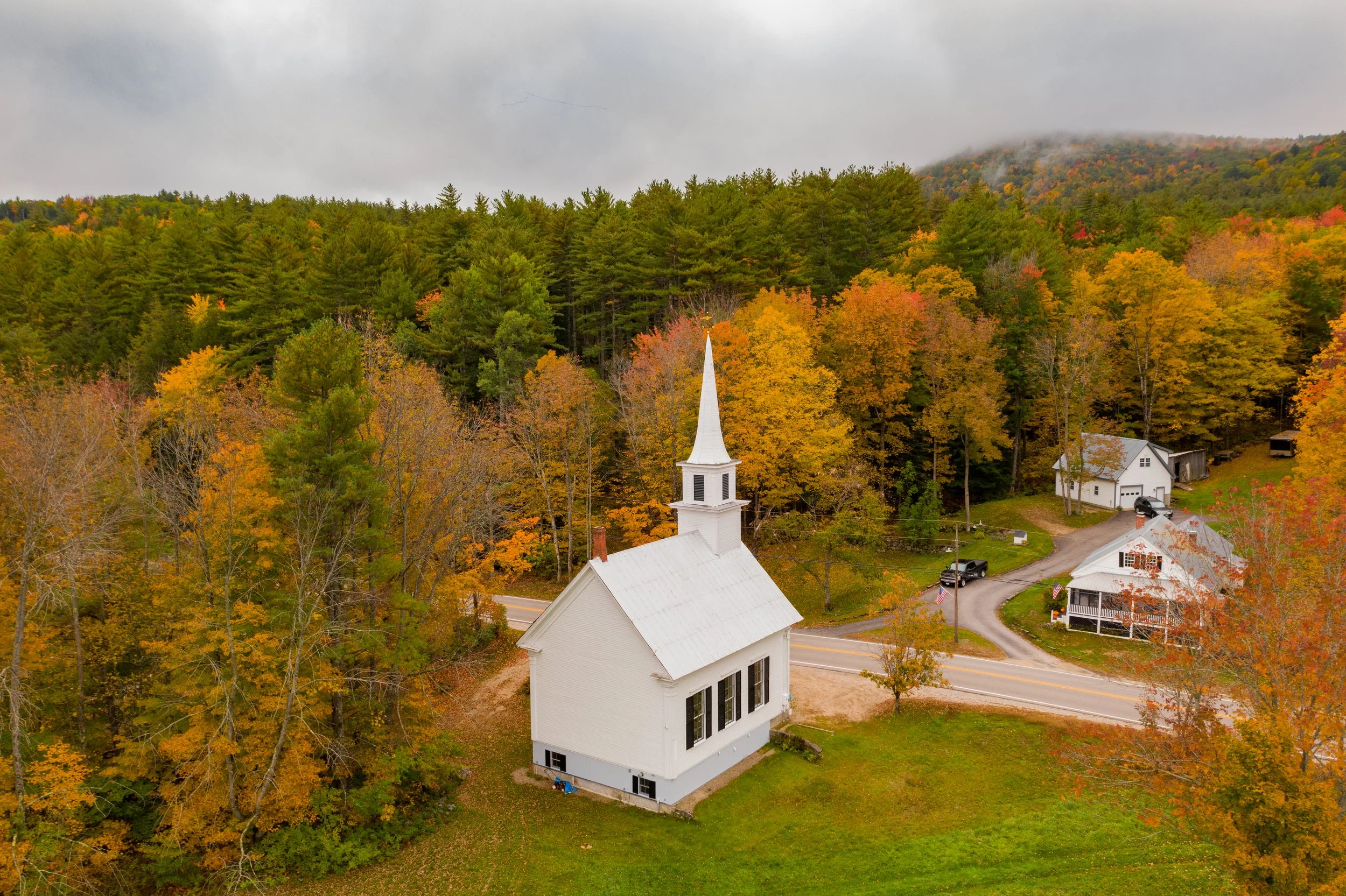 A white church with a tall steeple surrounded by autumn trees with colorful fall foliage in a rural setting, with mountains and overcast skies in the background.