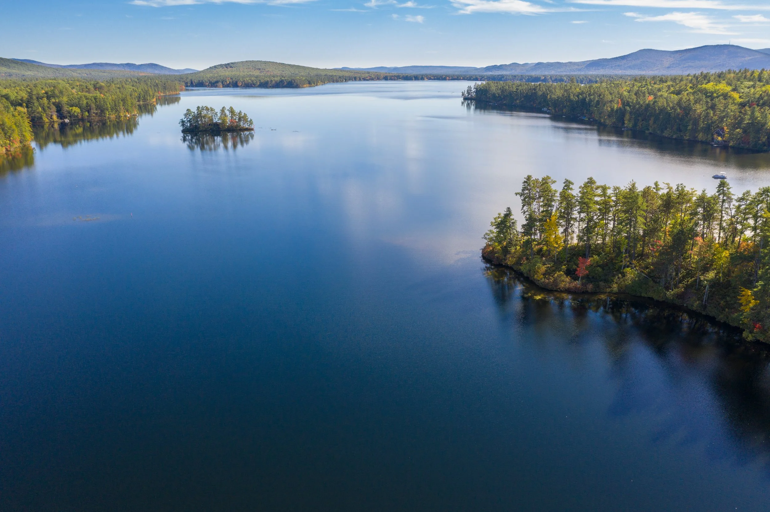 Aerial view of a large lake surrounded by dense forested hills and mountains, with clear blue water and a small boat near the shoreline.