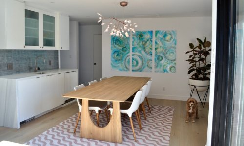 Modern dining room with a wooden table, white chairs, abstract wall art, and a dog near a potted plant. The room features a minimalist design with white cabinetry and green accents.